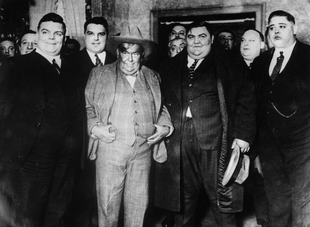 A group of well-dressed men pose together at a formal event. The central figure wears a suit and hat, surrounded by others in suits