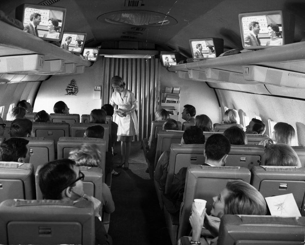 Flight attendants serve passengers in a vintage airplane cabin with overhead screens playing a show