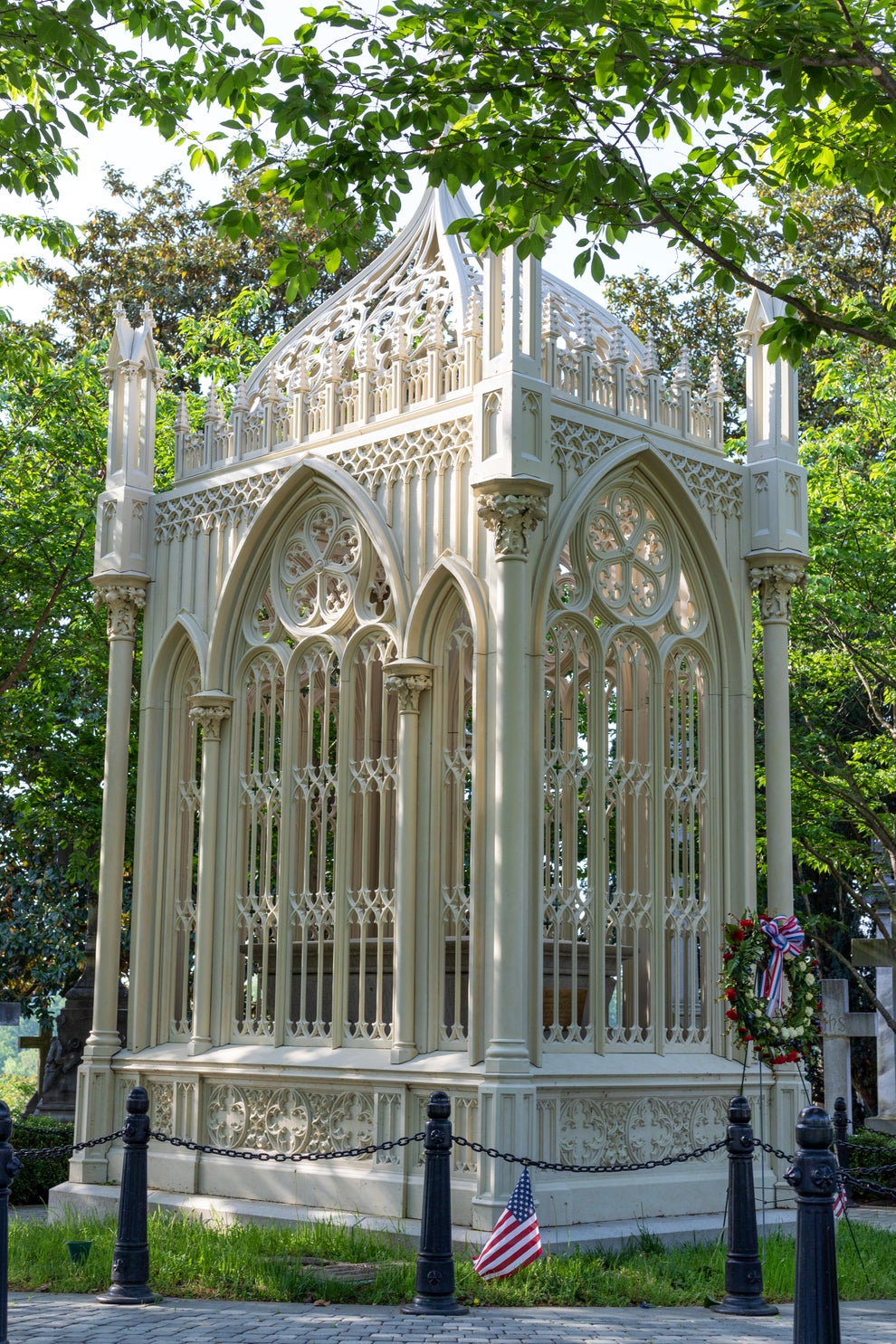 Ornate Gothic-style gazebo with arched windows and intricate designs, surrounded by greenery and a small flag at the base