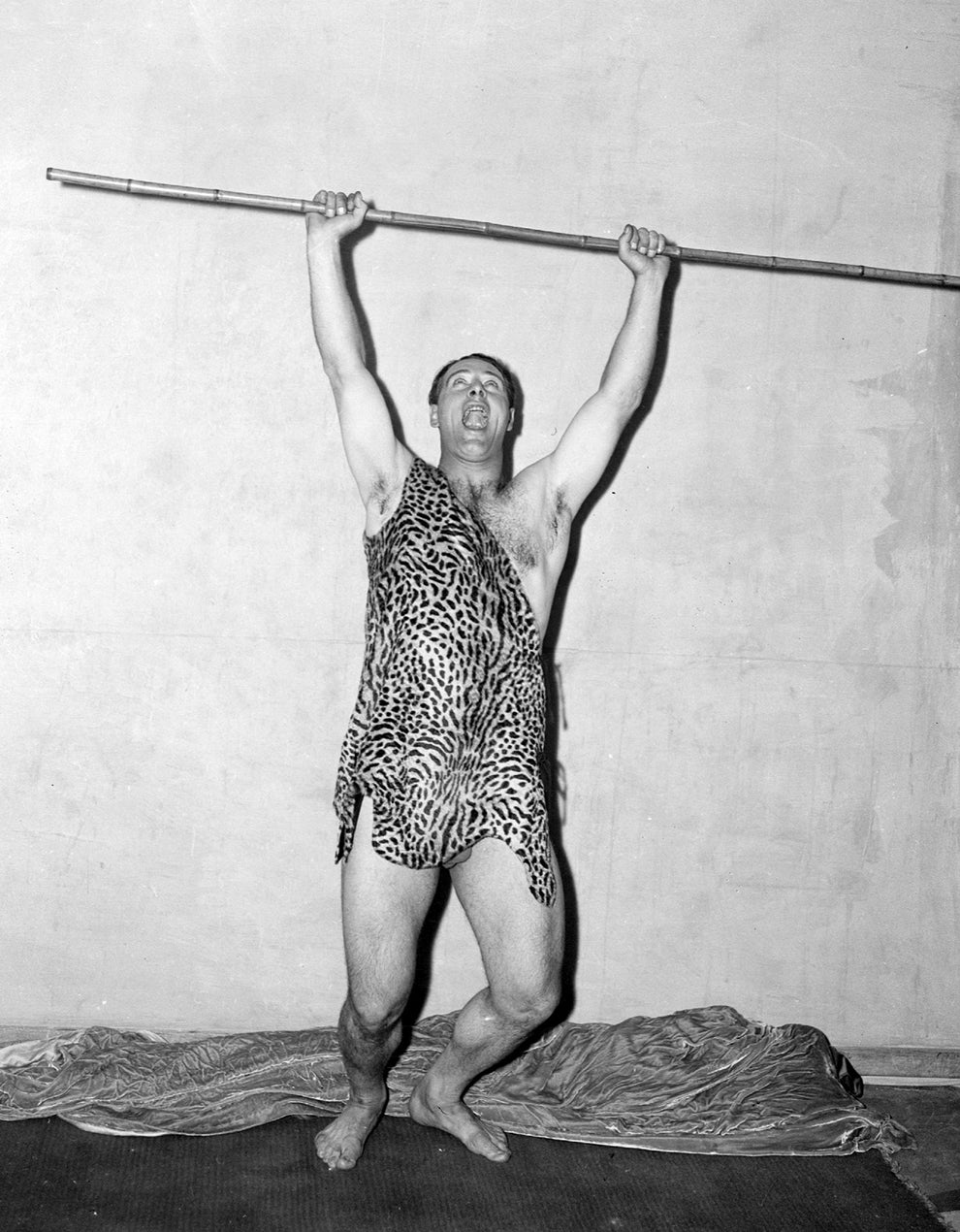 Person posing joyfully while holding a pole overhead, wearing a leopard-print tunic resembling a classic Tarzan costume