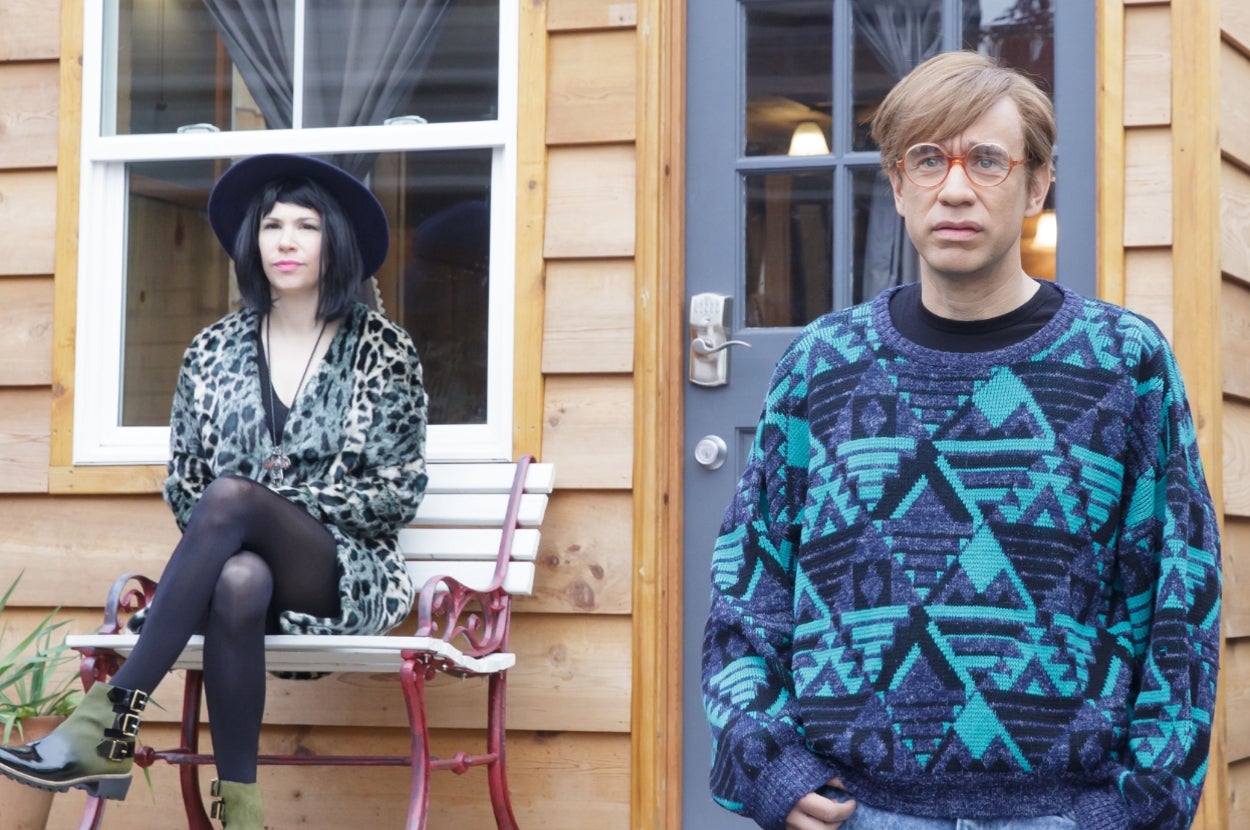 Two individuals pose outside a wooden cabin; one wears a patterned sweater and glasses, the other a leopard-print coat and wide-brim hat