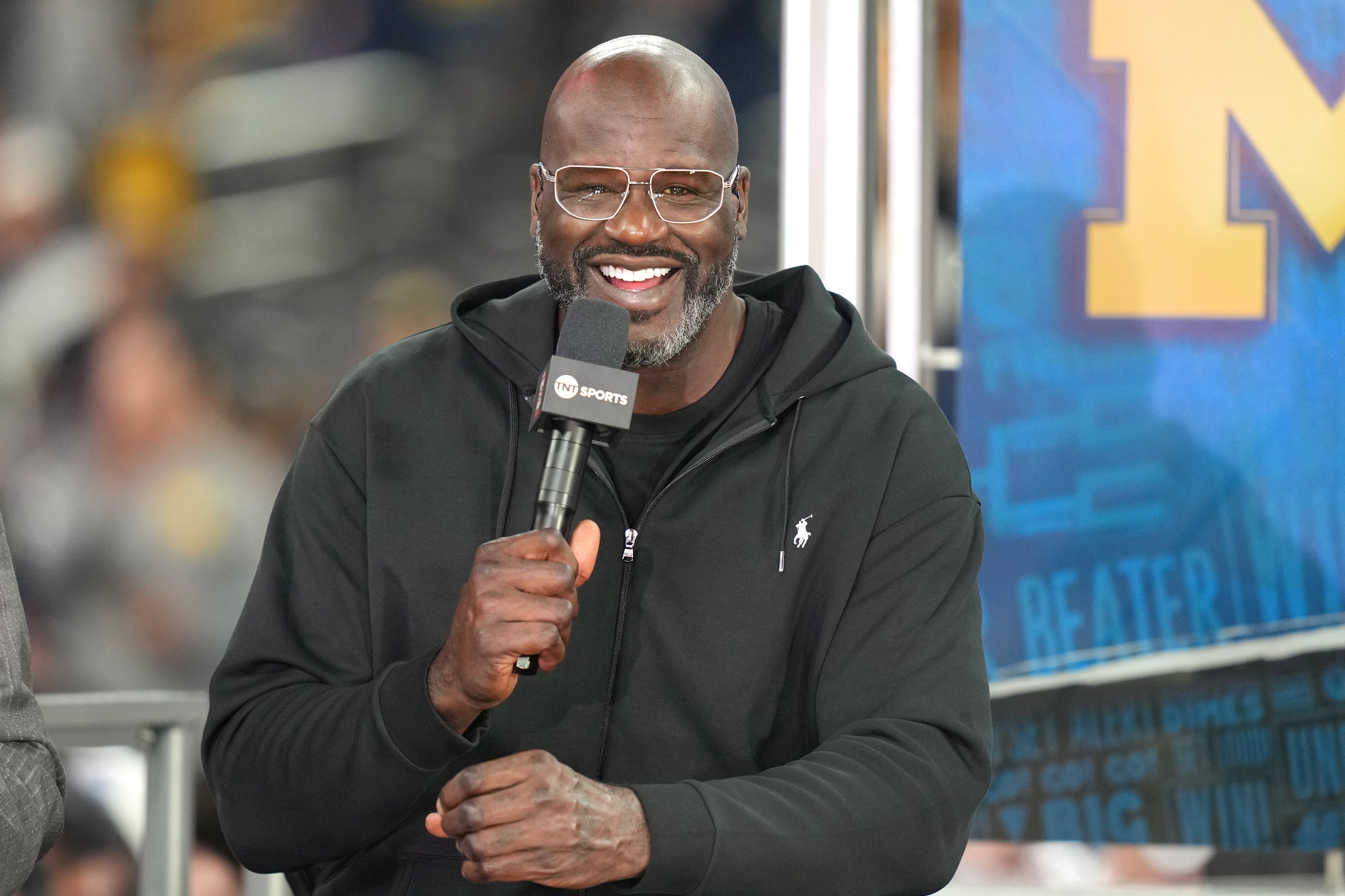 Person in a casual black hoodie holds a microphone, smiling, at a sports event