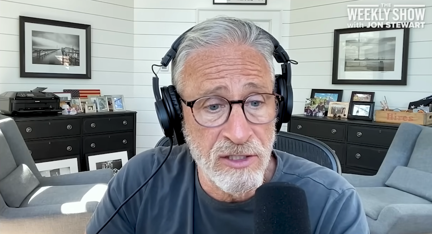 En person med hodetelefoner og briller snakker inn i en mikrofon under en podcast eller videosamtale, omgitt av innrammede bilder og dekor