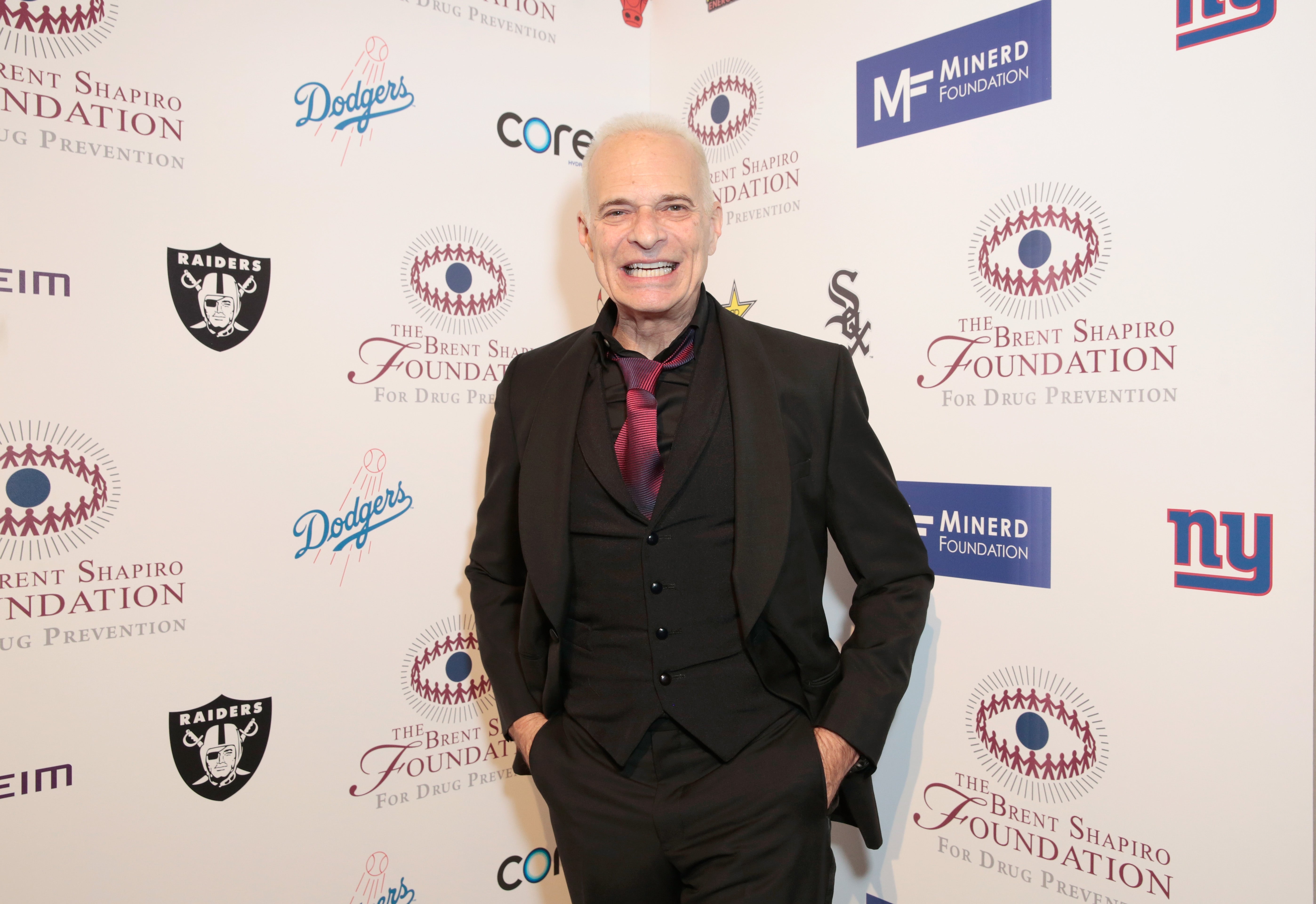 Person in a black suit and shirt with a red tie at a charity event backdrop featuring various brand logos