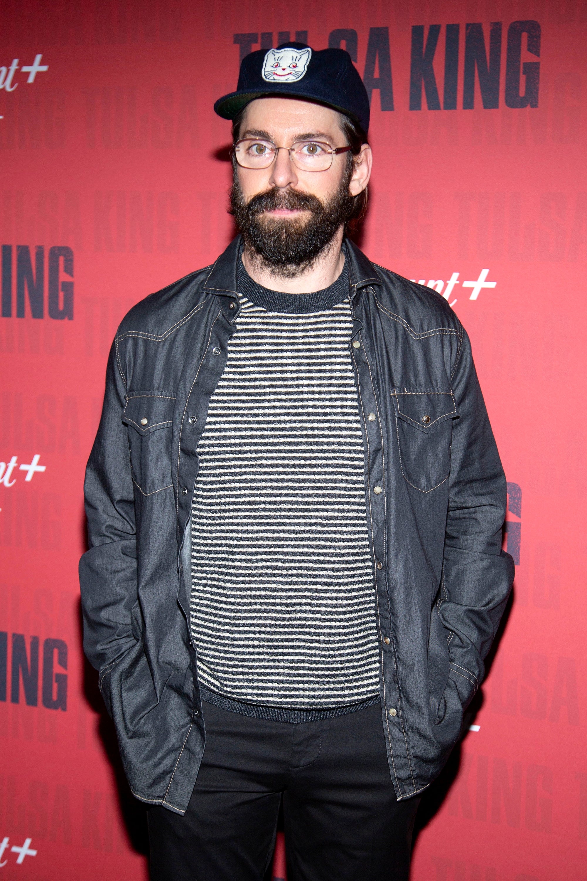 Person on red carpet event, wearing a casual striped shirt, denim jacket, and cap, posing for cameras against a backdrop with "The SA King" text