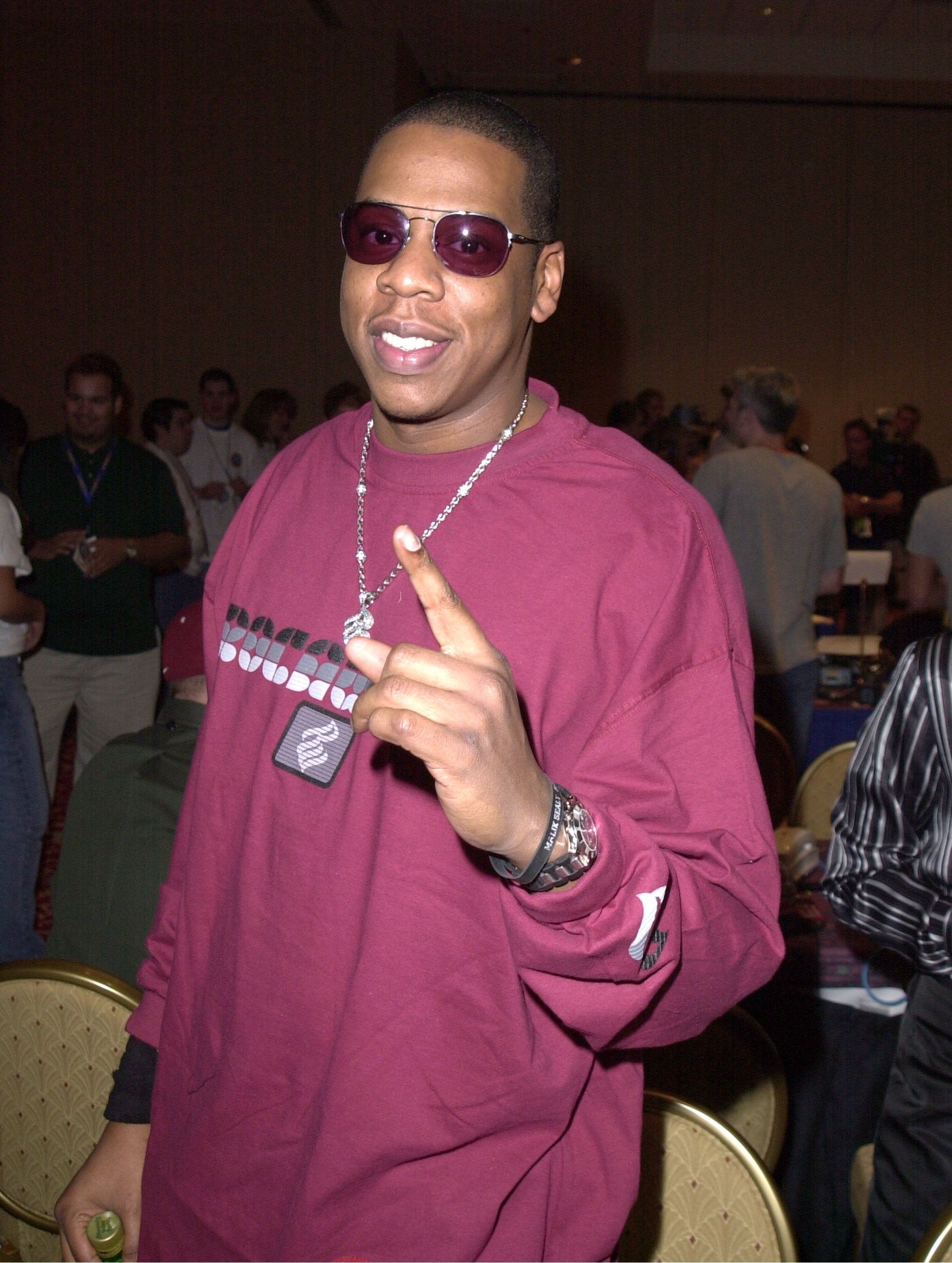 Person wearing sunglasses and casual baggy attire, gesturing with finger up, in a social gathering setting