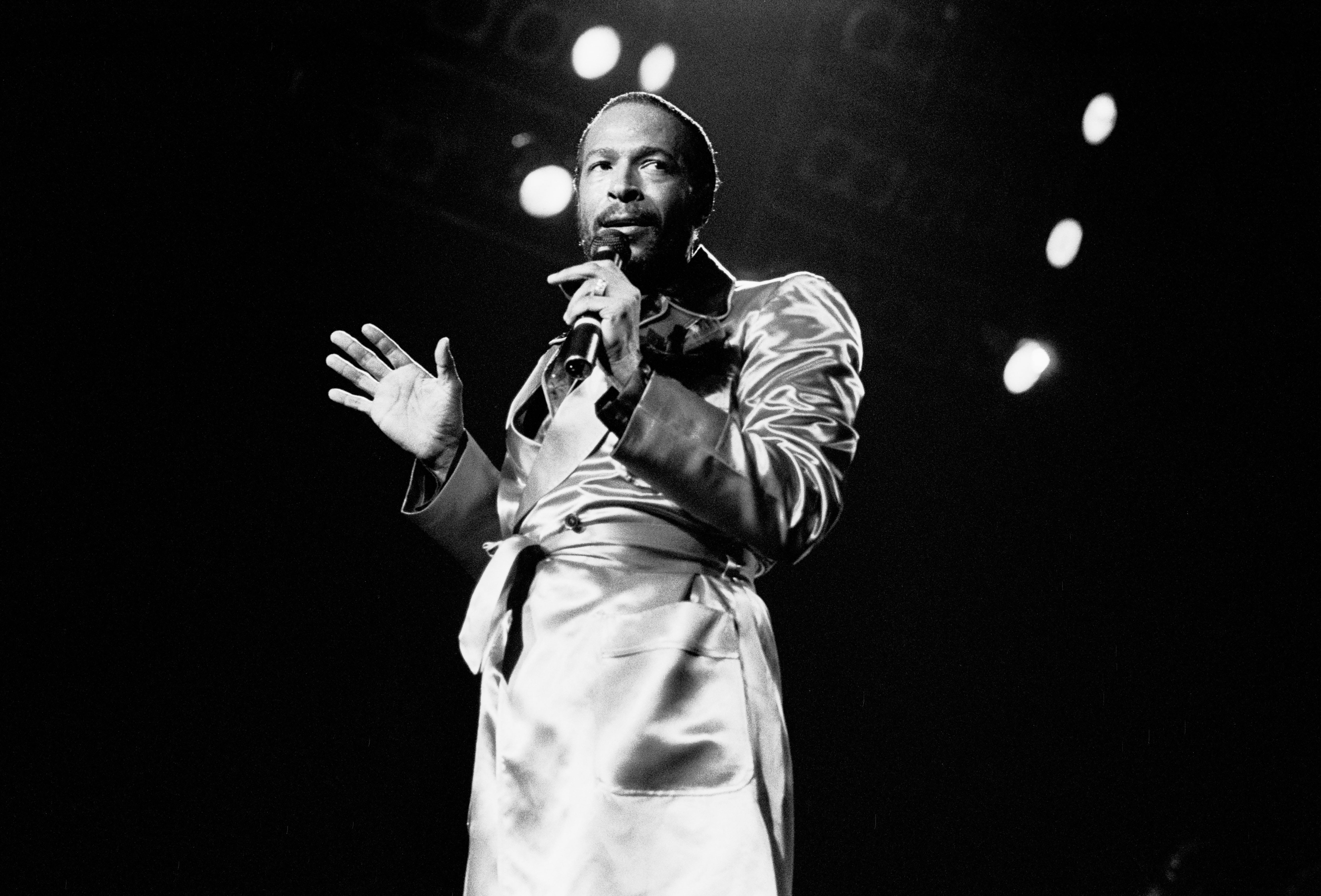 Singer on stage wearing a stylish satin suit, holding a microphone. Stage lights shine behind, creating a dramatic scene
