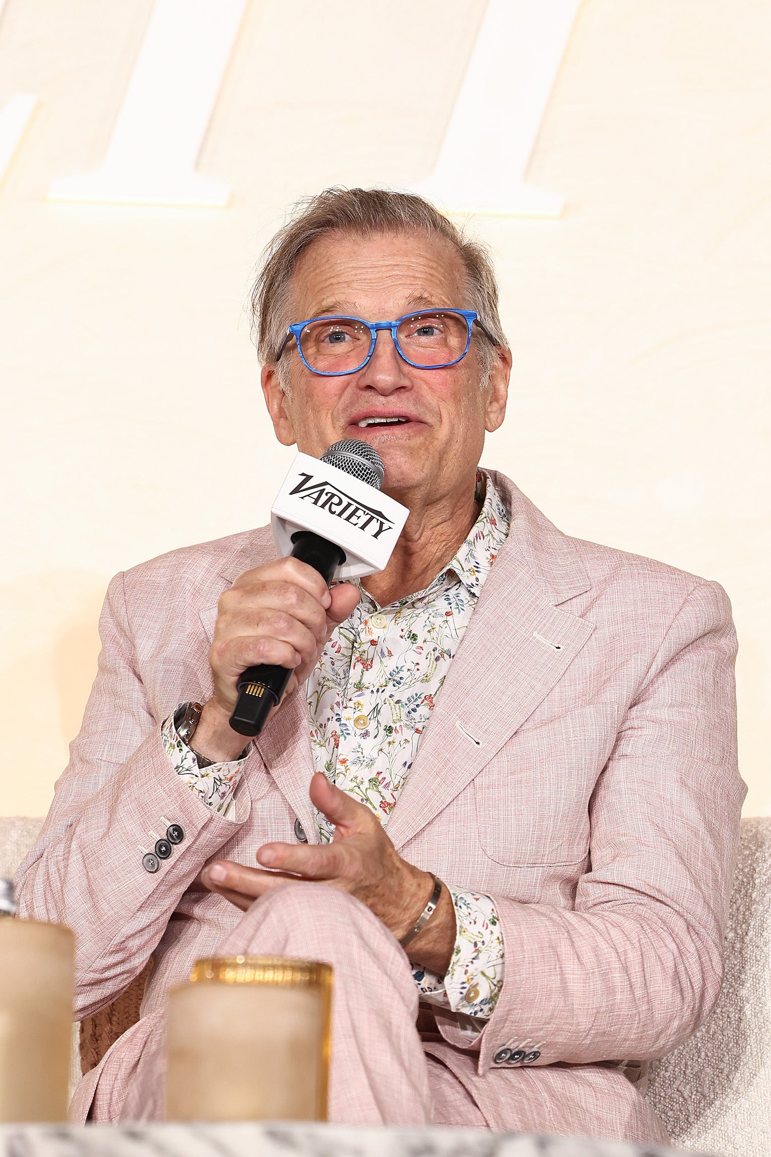 Person speaking at an event, wearing a light patterned suit and floral shirt, holding a microphone labeled "Variety."