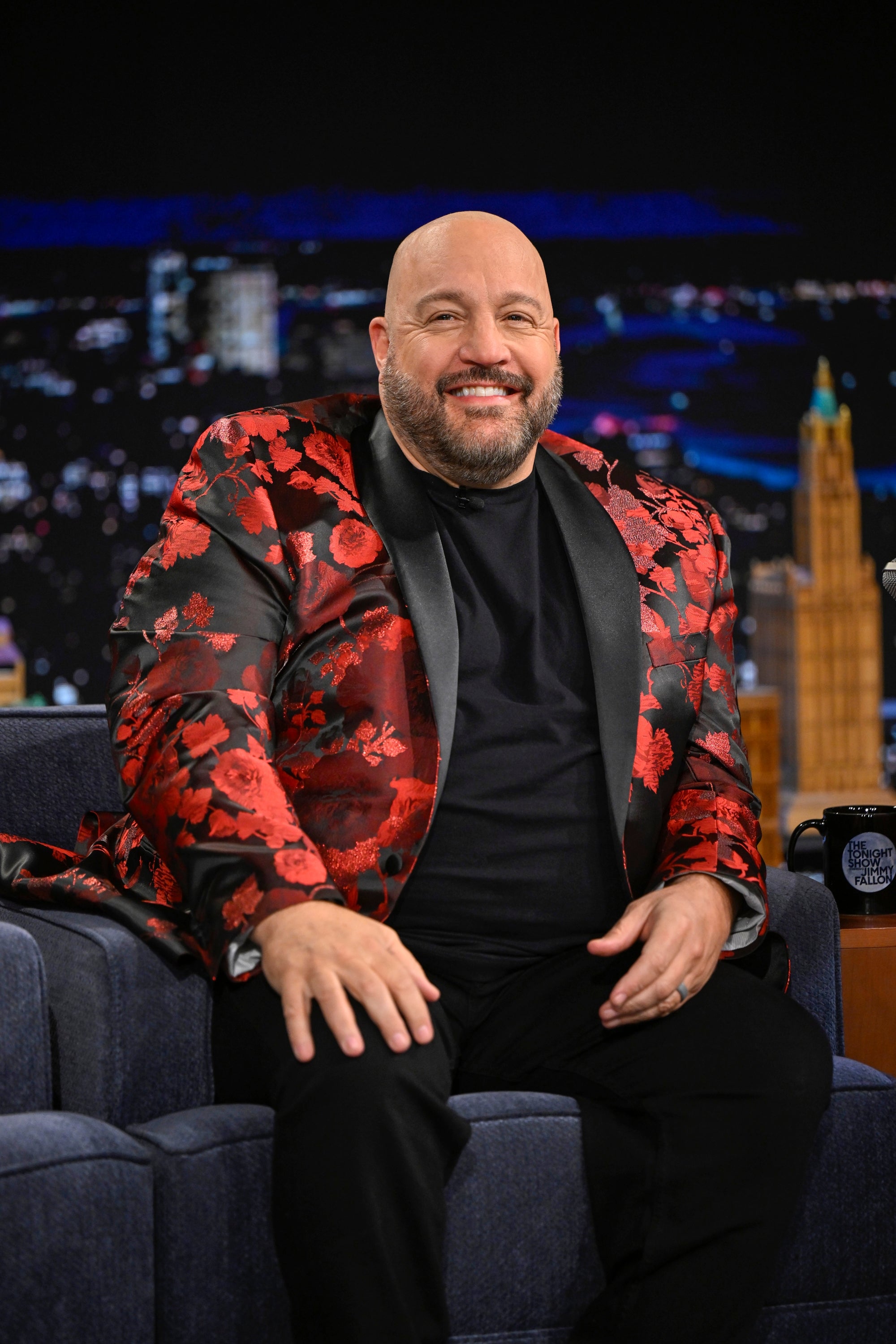 Person seated on a talk show set, wearing a floral-patterned jacket and a black shirt, smiling for the camera