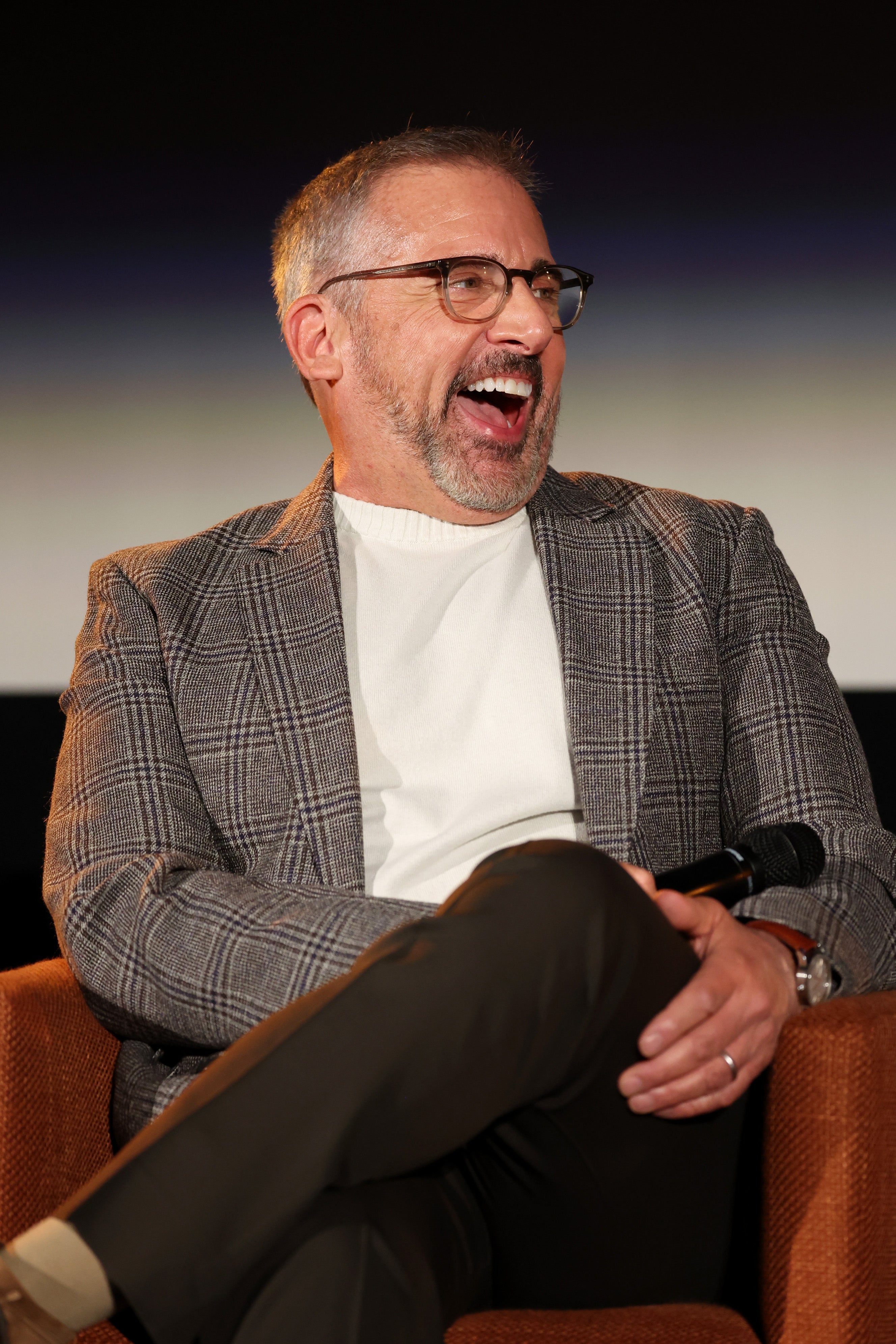 A man in a plaid blazer and glasses laughs while sitting, conveying a relaxed, joyful demeanor at a work-related event