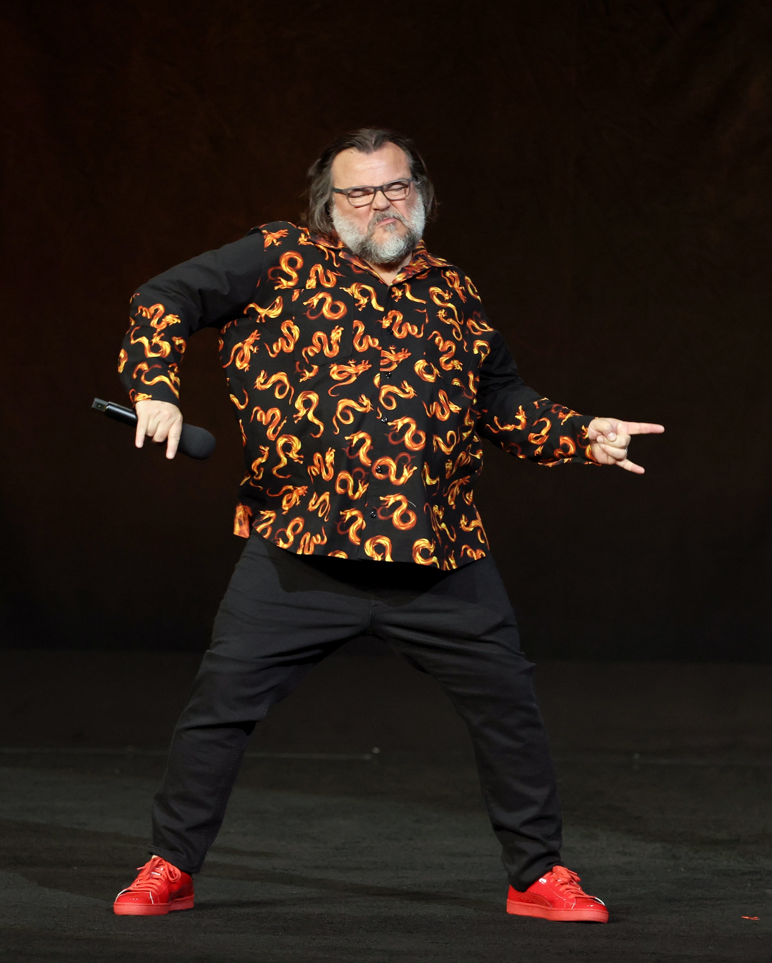 Person on stage striking a playful pose, wearing a patterned shirt and red shoes, holding a microphone