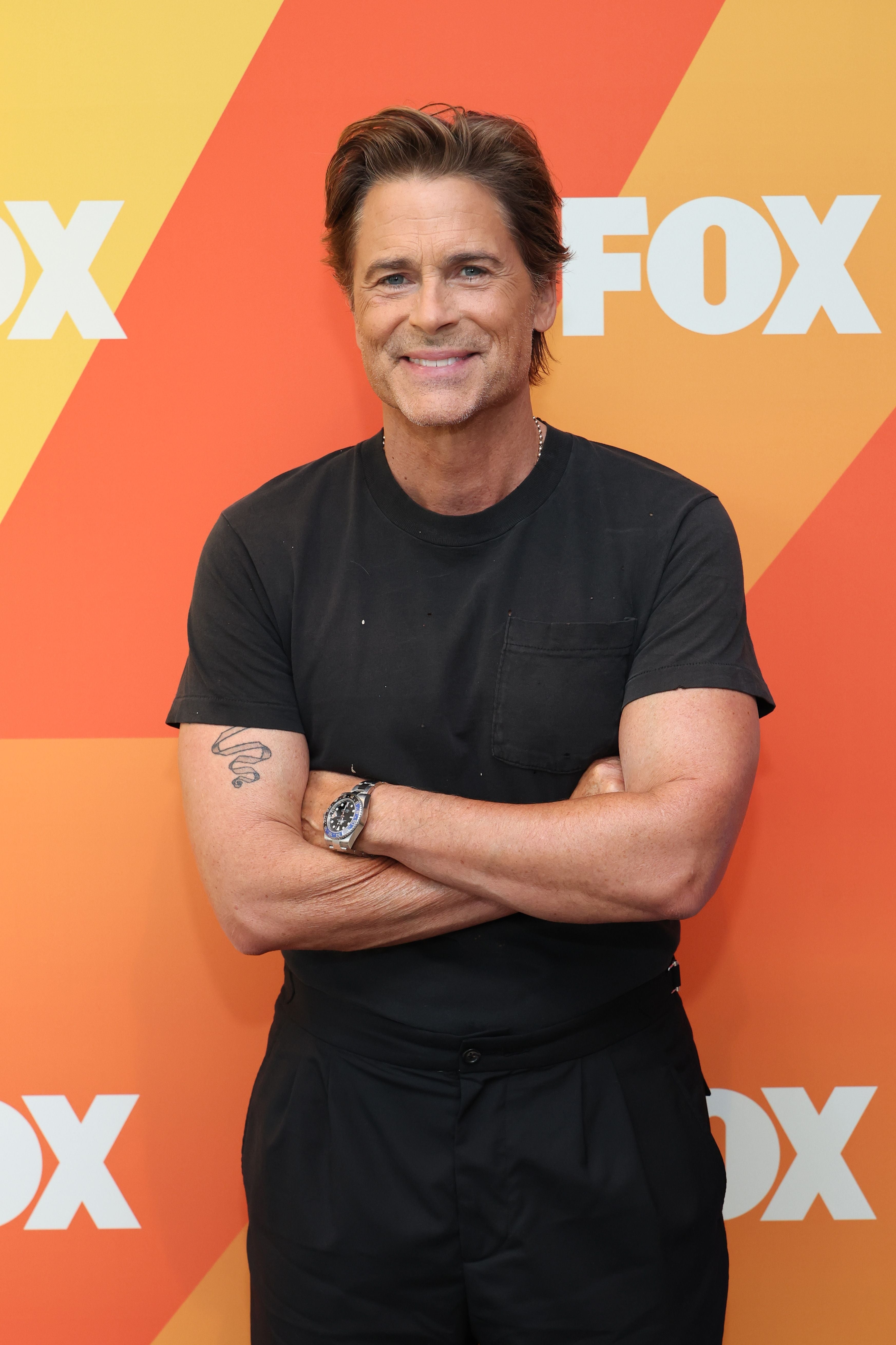 Smiling man in a black t-shirt with arms crossed at a FOX event backdrop, suggesting a casual yet professional appearance