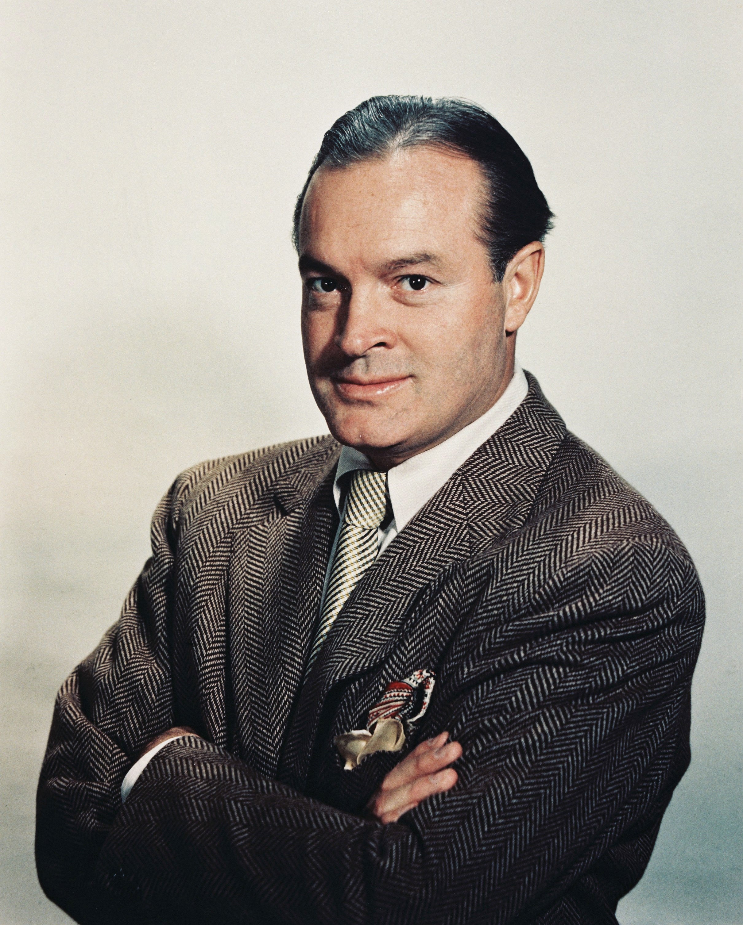 Person in a herringbone suit jacket and tie, arms crossed, looking confidently at the camera