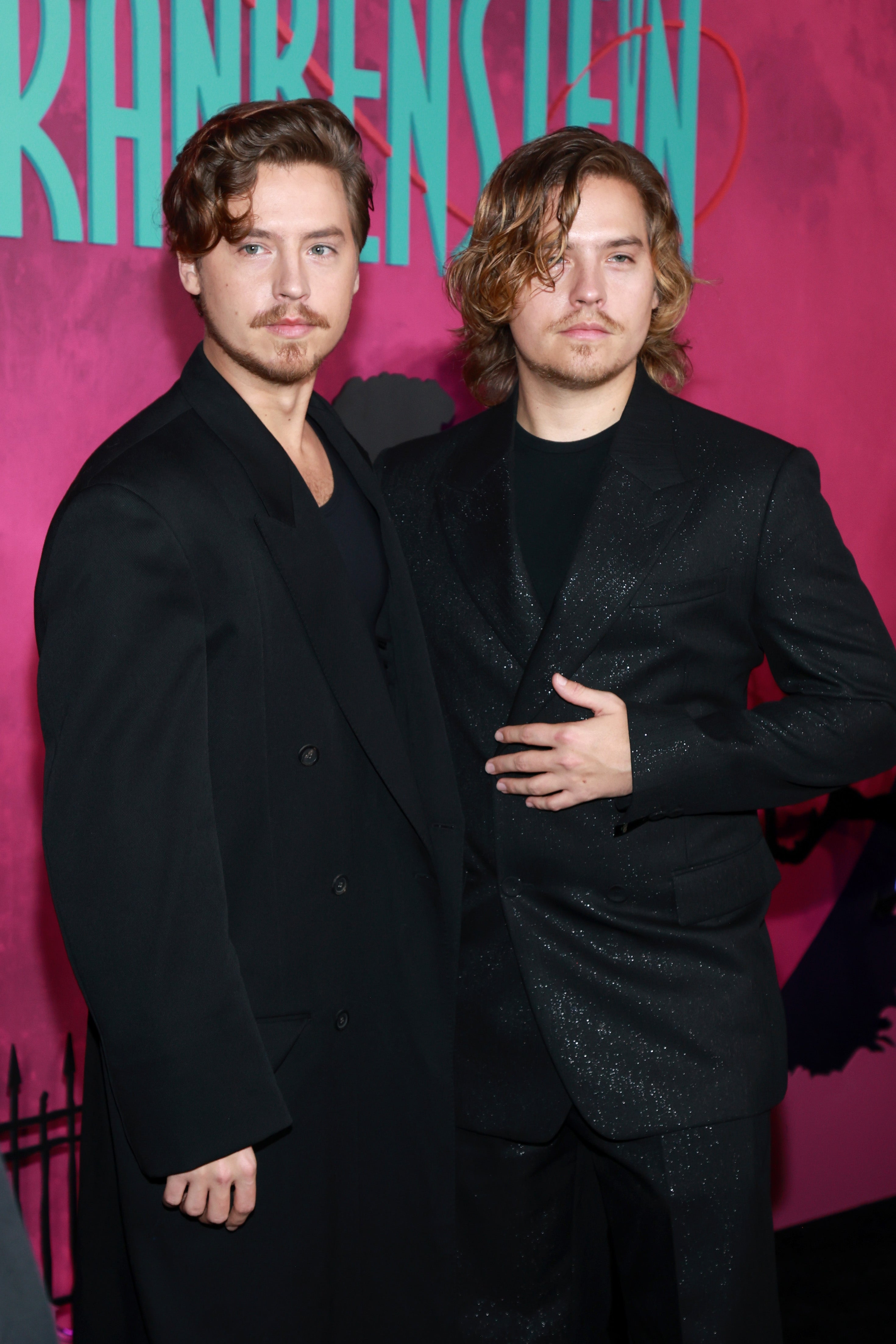 Two individuals in black suits pose at an event backdrop