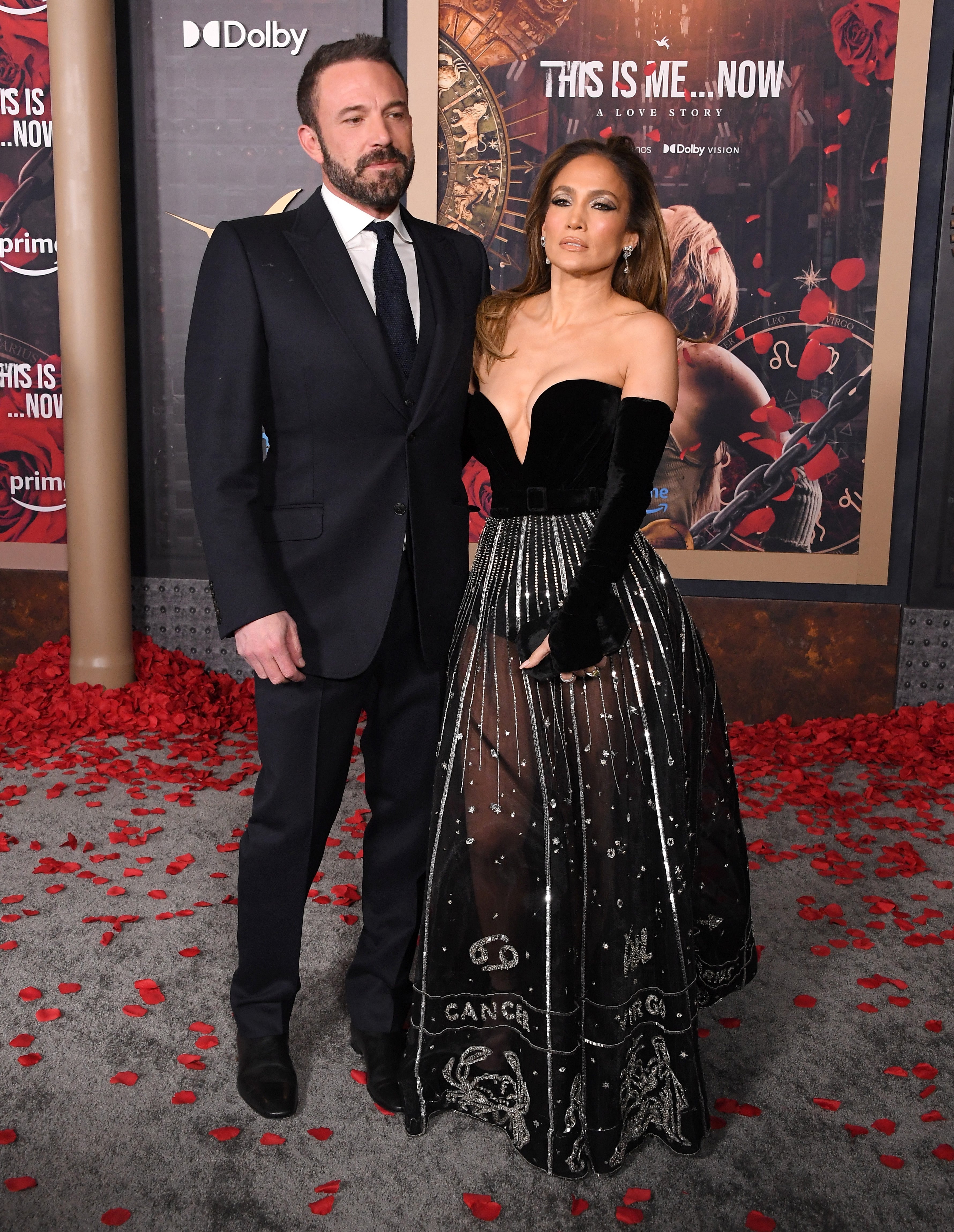 Two individuals on a red carpet event. One wears a long, strapless black gown with intricate embellishments; the other wears a dark suit