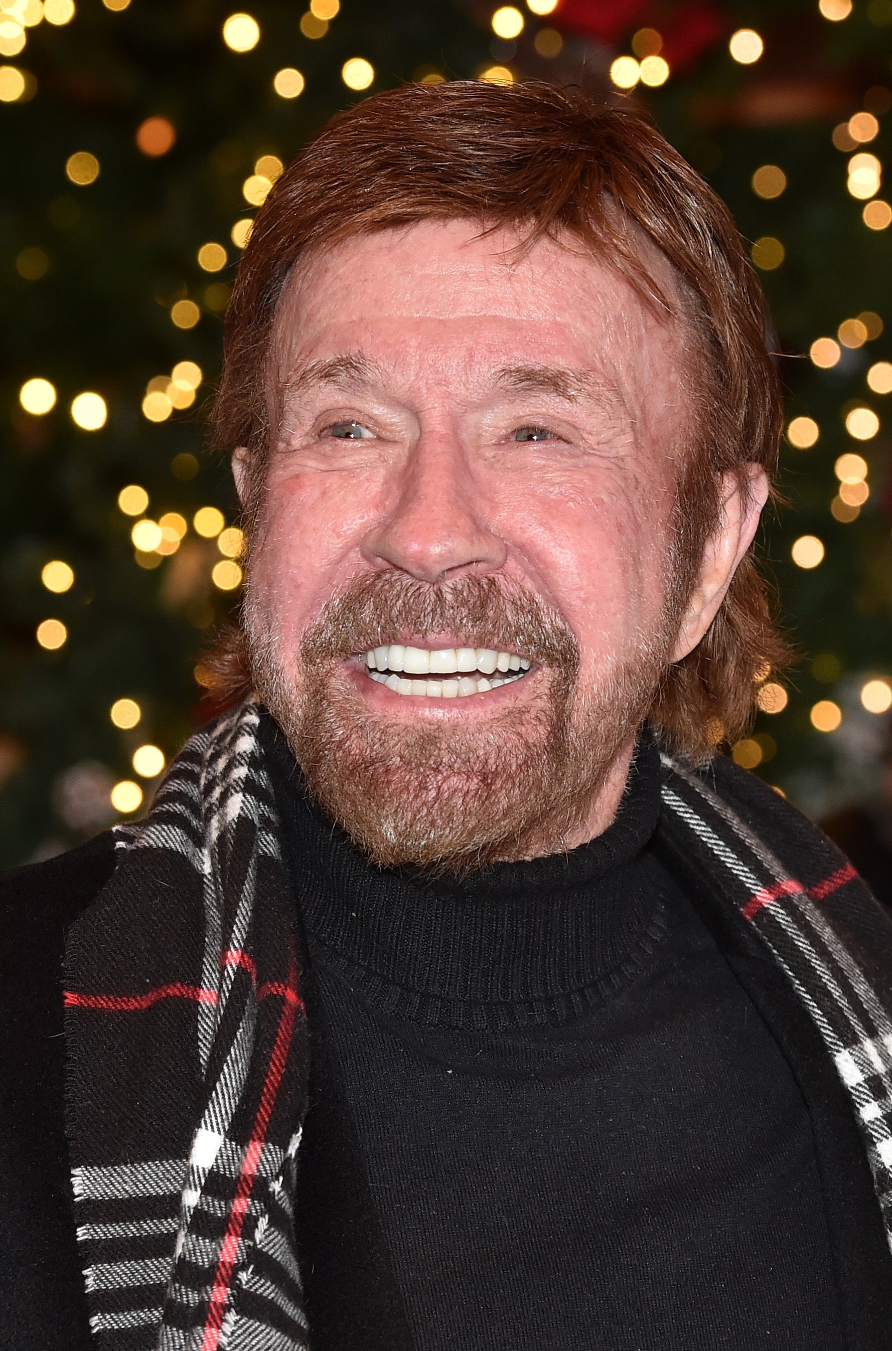 A person smiling broadly, wearing a black turtleneck and plaid scarf, stands in front of a festive backdrop with twinkling lights