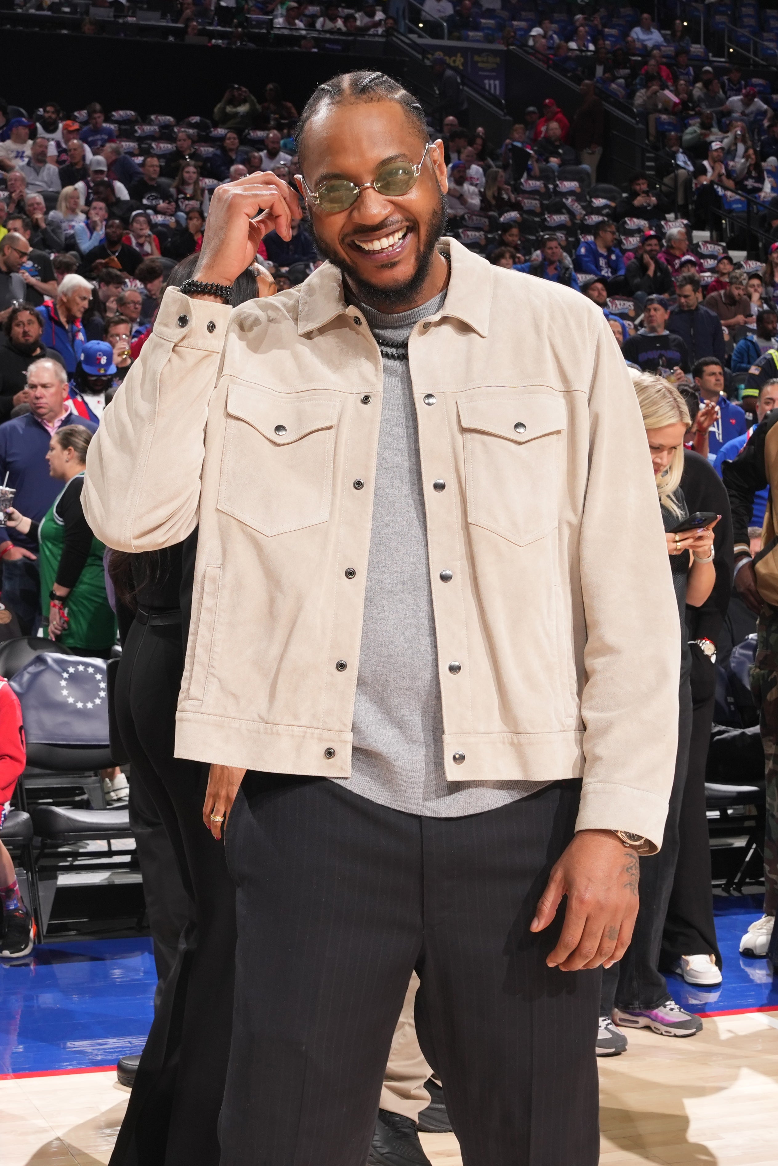 Person smiling at a sports event, wearing a casual jacket, sweater, and pants. They engage with the crowd, suggesting a relaxed, approachable demeanor