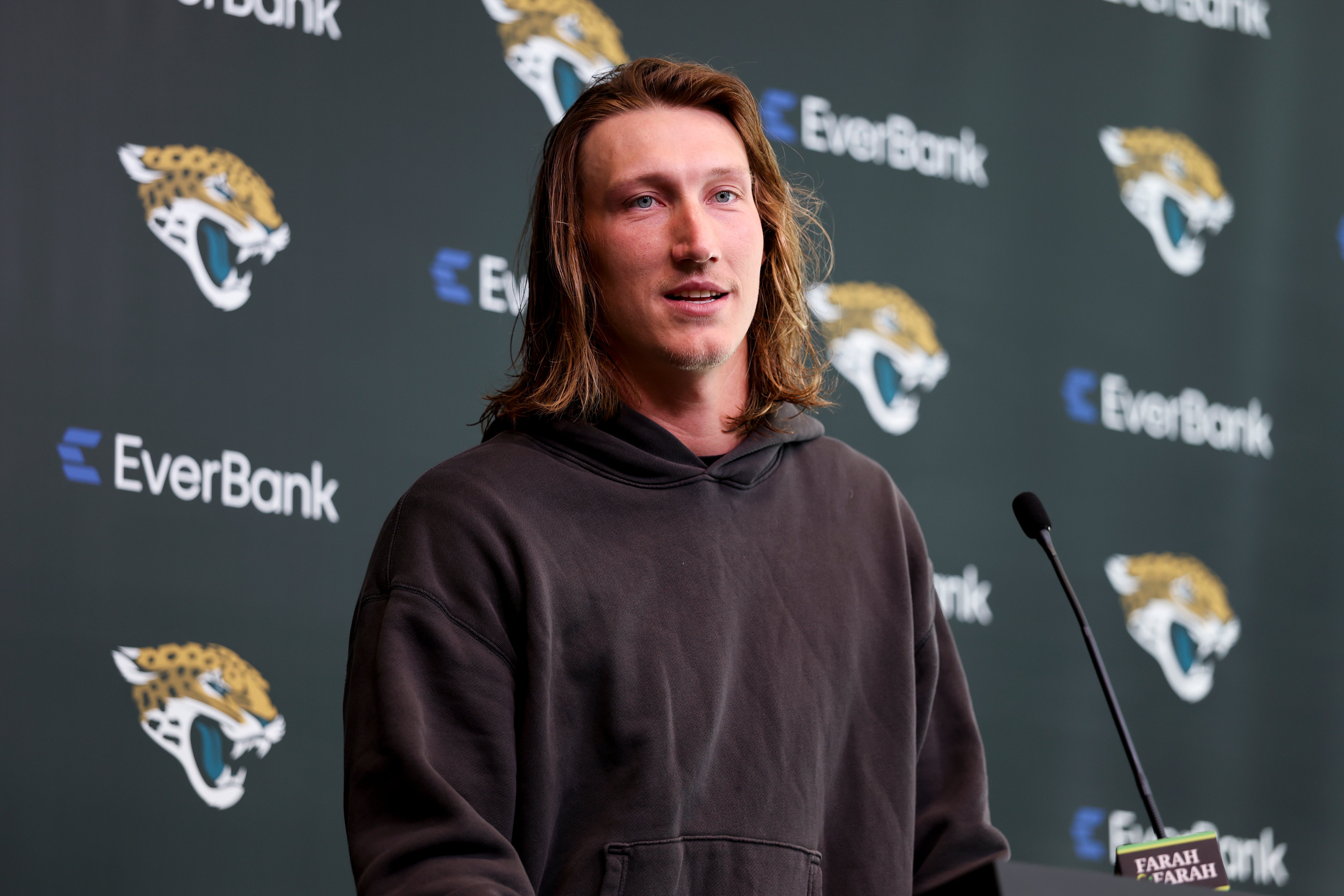 Person speaking at podium with Jaguars logo backdrop, wearing a casual hoodie. Microphone is in the foreground