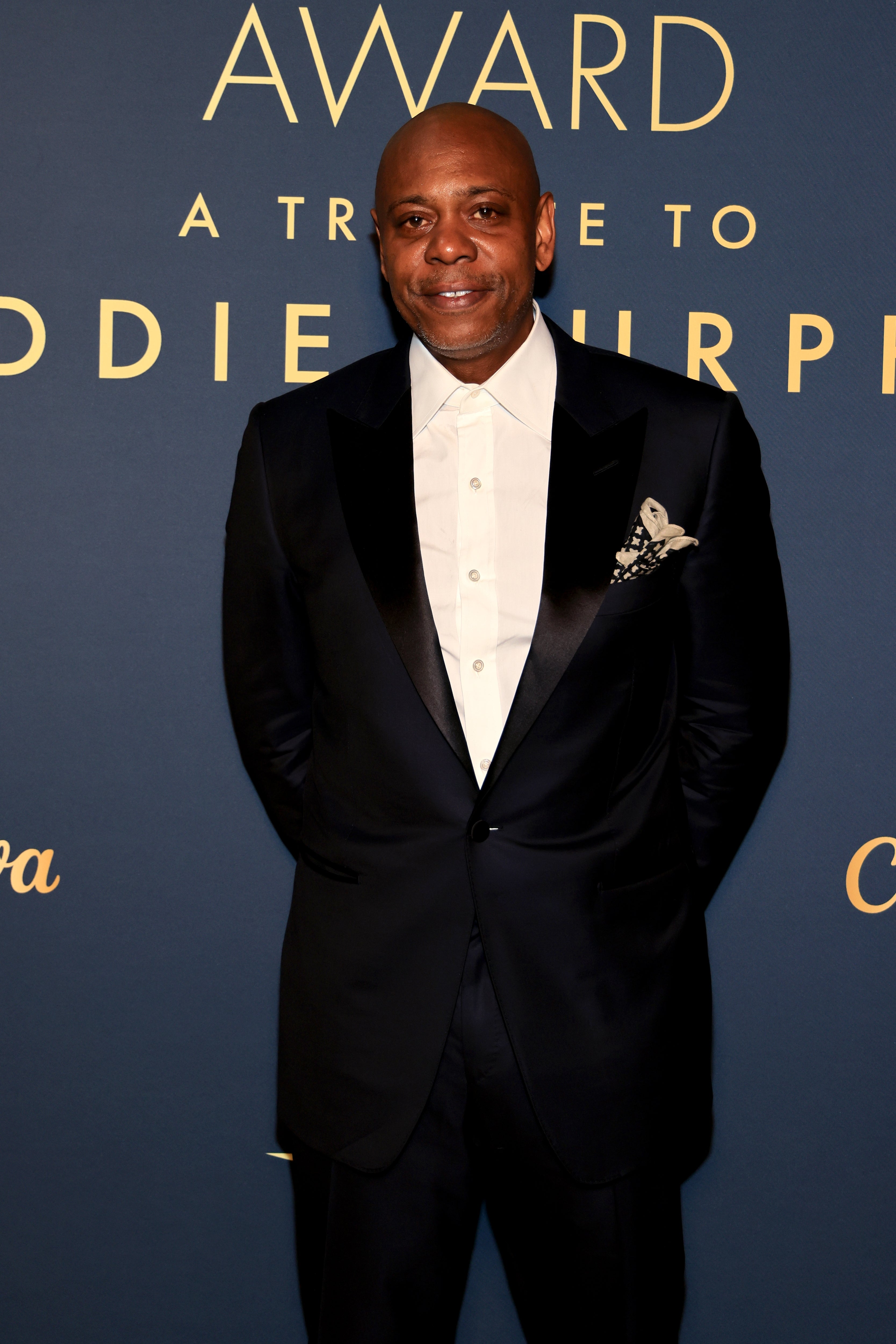 Person in a formal black suit with a white shirt stands in front of an event backdrop related to a tribute