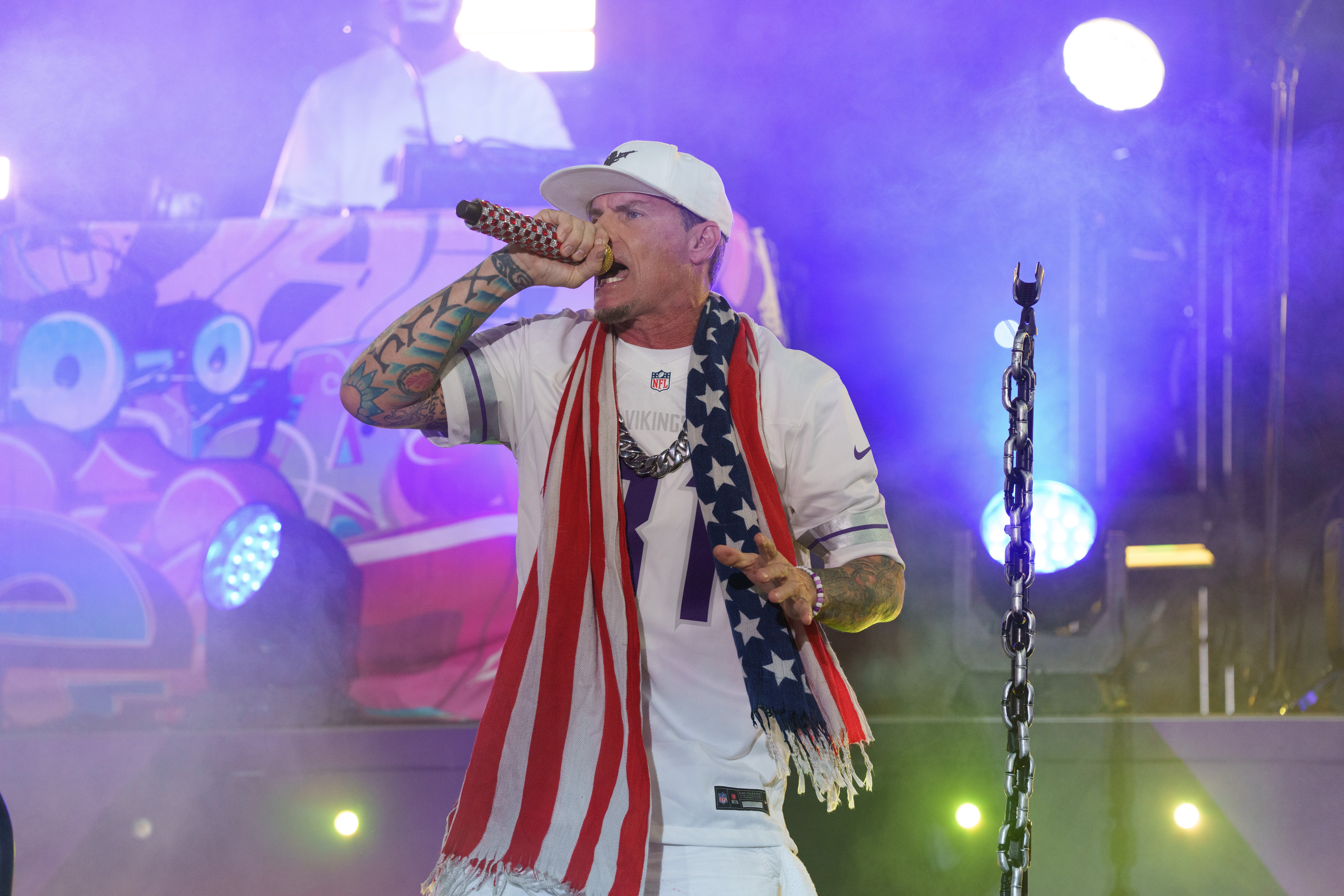 A performer connected  signifier    with a microphone, wearing a sports jersey, cap, and an American flag-themed scarf, successful  beforehand   of agleam  lights and a DJ