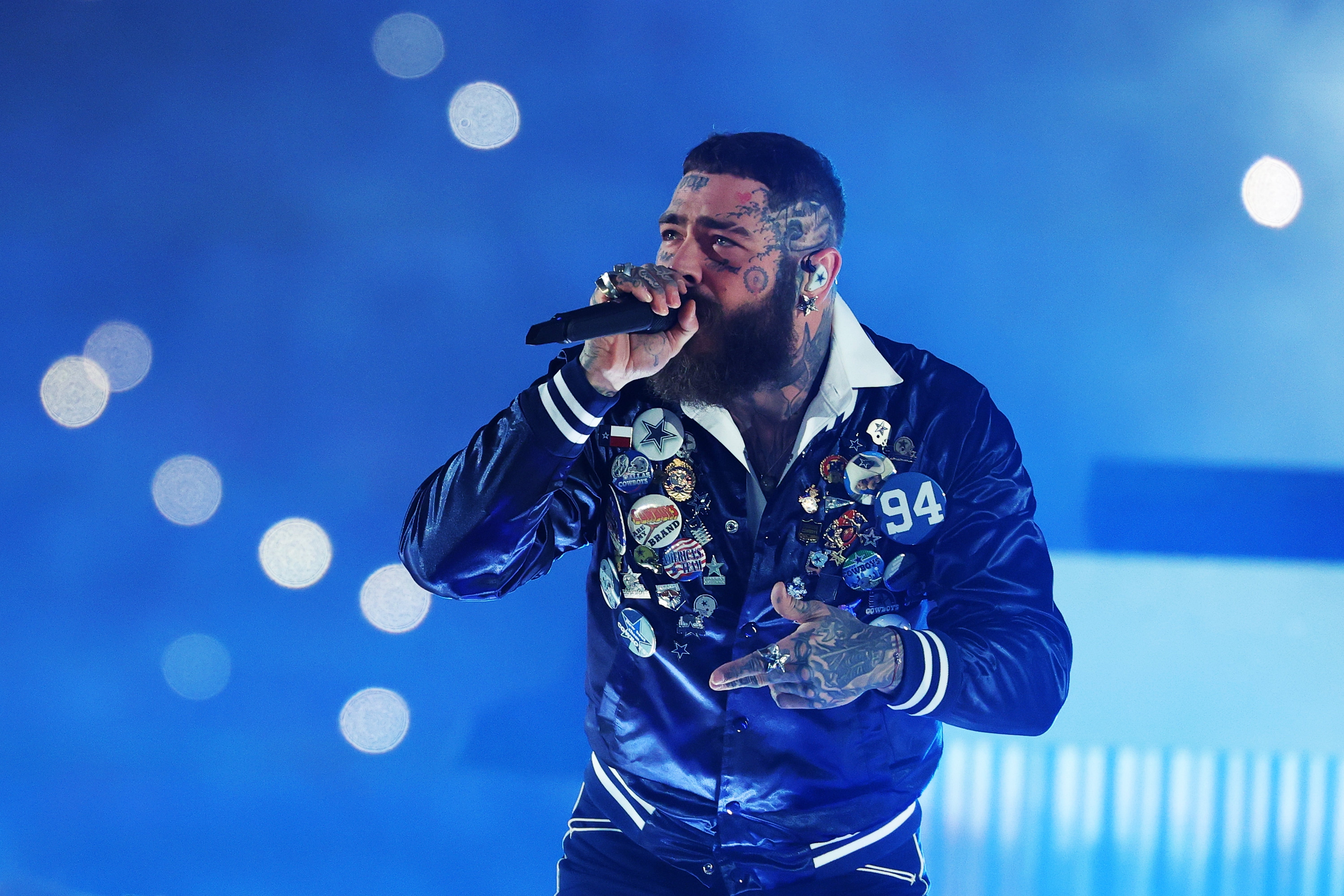 Musician performing on stage, wearing a jacket adorned with various patches, holding a microphone