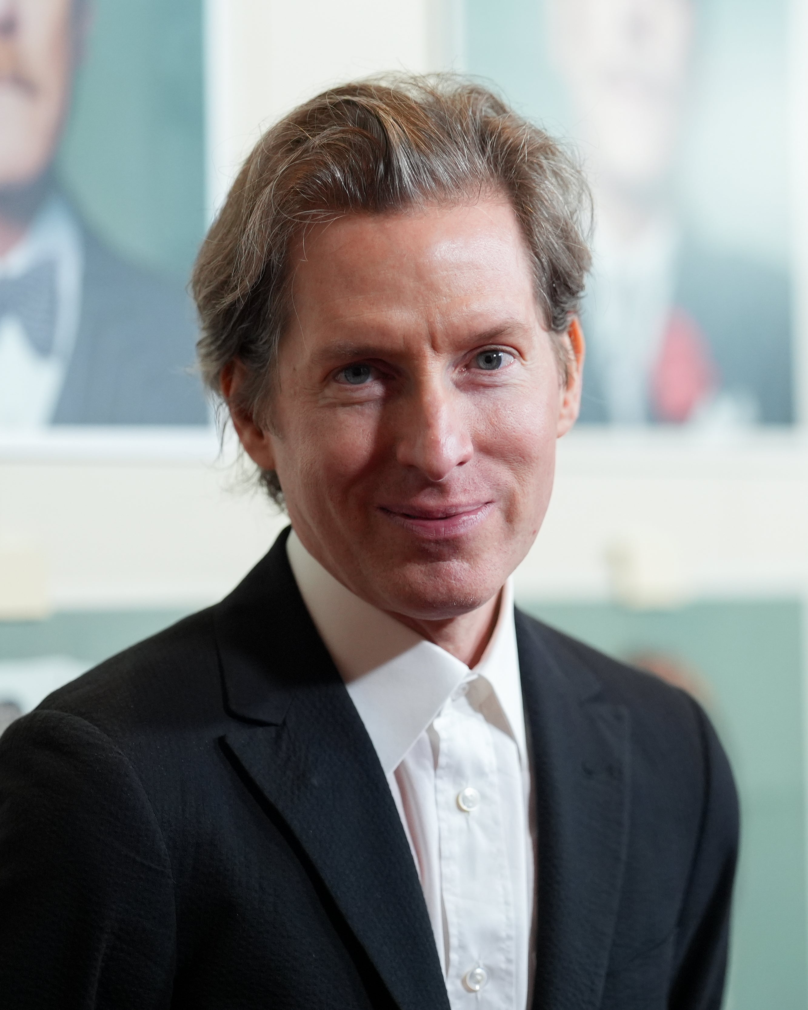 A person in a tailored suit smiles at the camera, standing in front of portrait-style photos