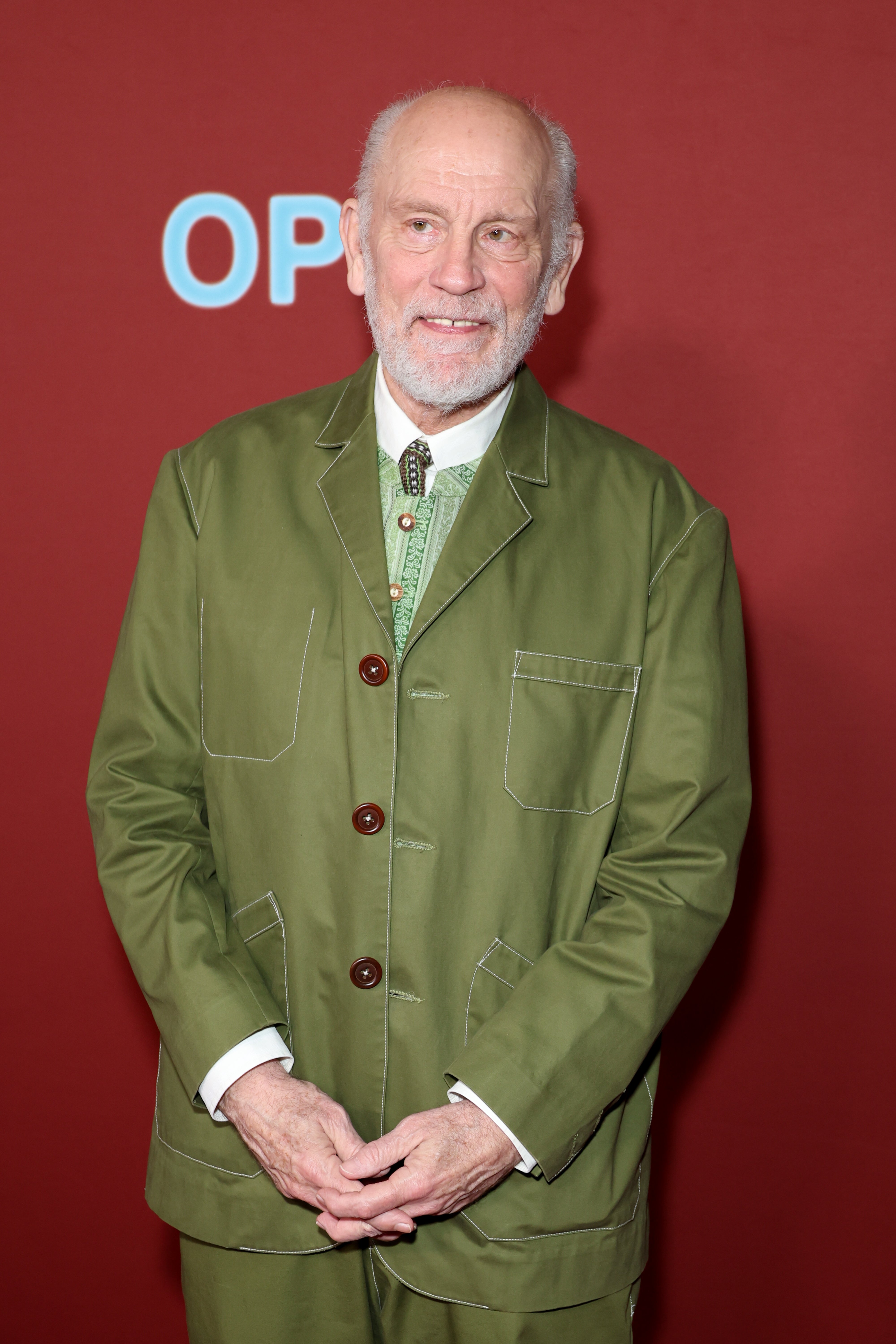 Person in a green suit with a white shirt on a red carpet at a media event, standing in front of a red background