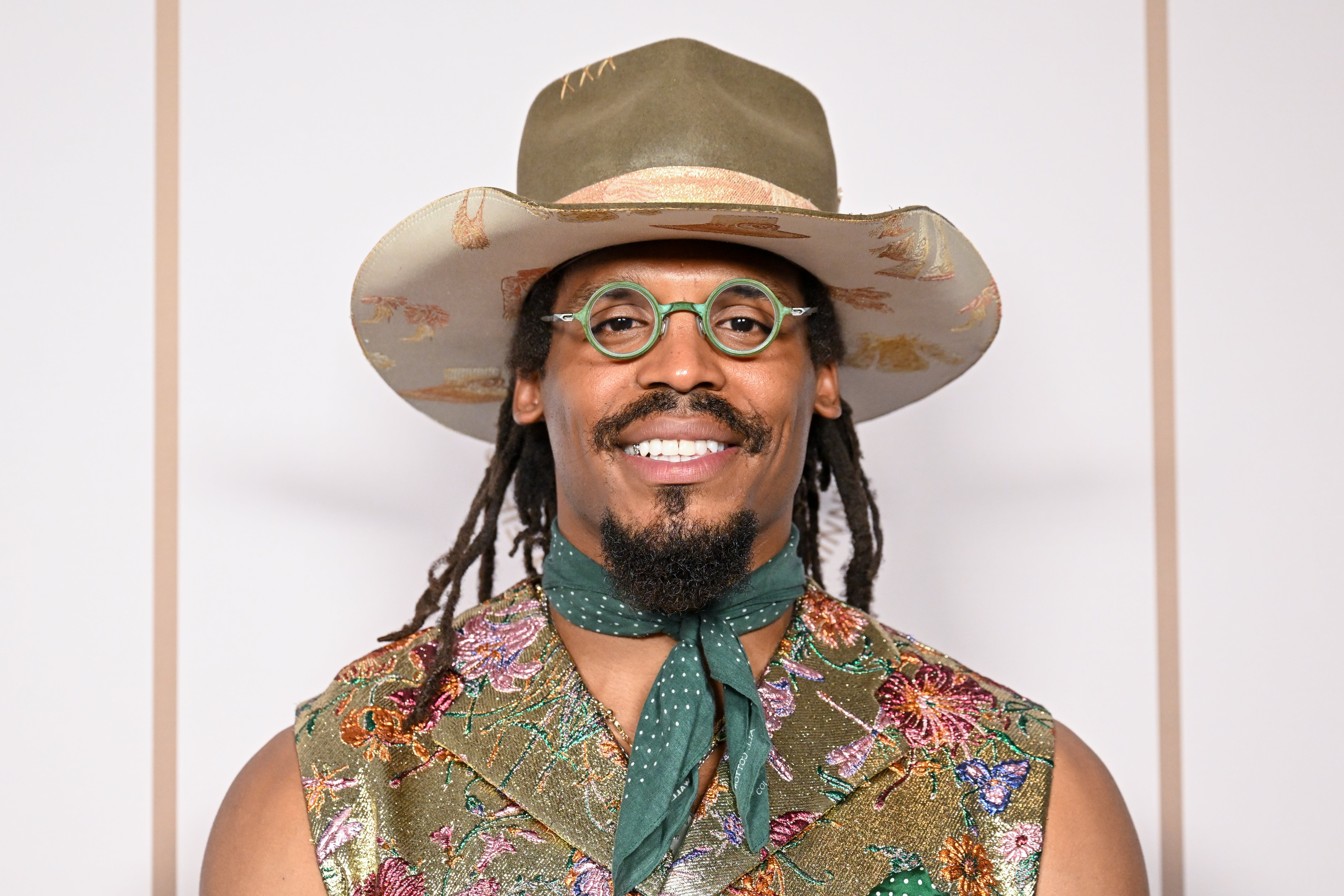 Person wearing a wide-brimmed hat and floral-patterned vest with a green scarf. They have round glasses and braided hair