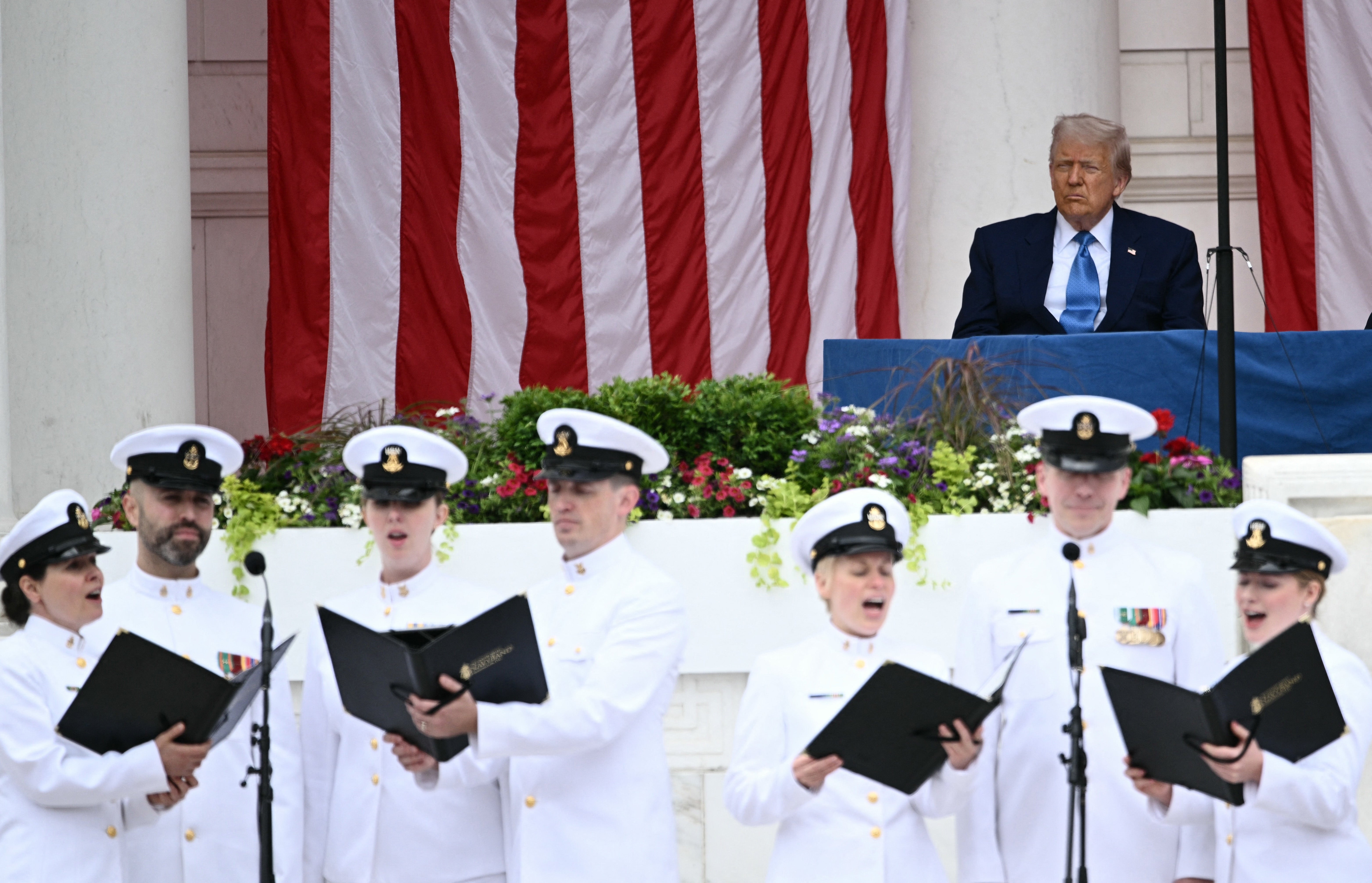 A radical  of naval officers successful  achromatic  uniforms sing astatine  a ceremony. A seated antheral   successful  a bluish  suit   watches from a podium adorned with flags and flowers