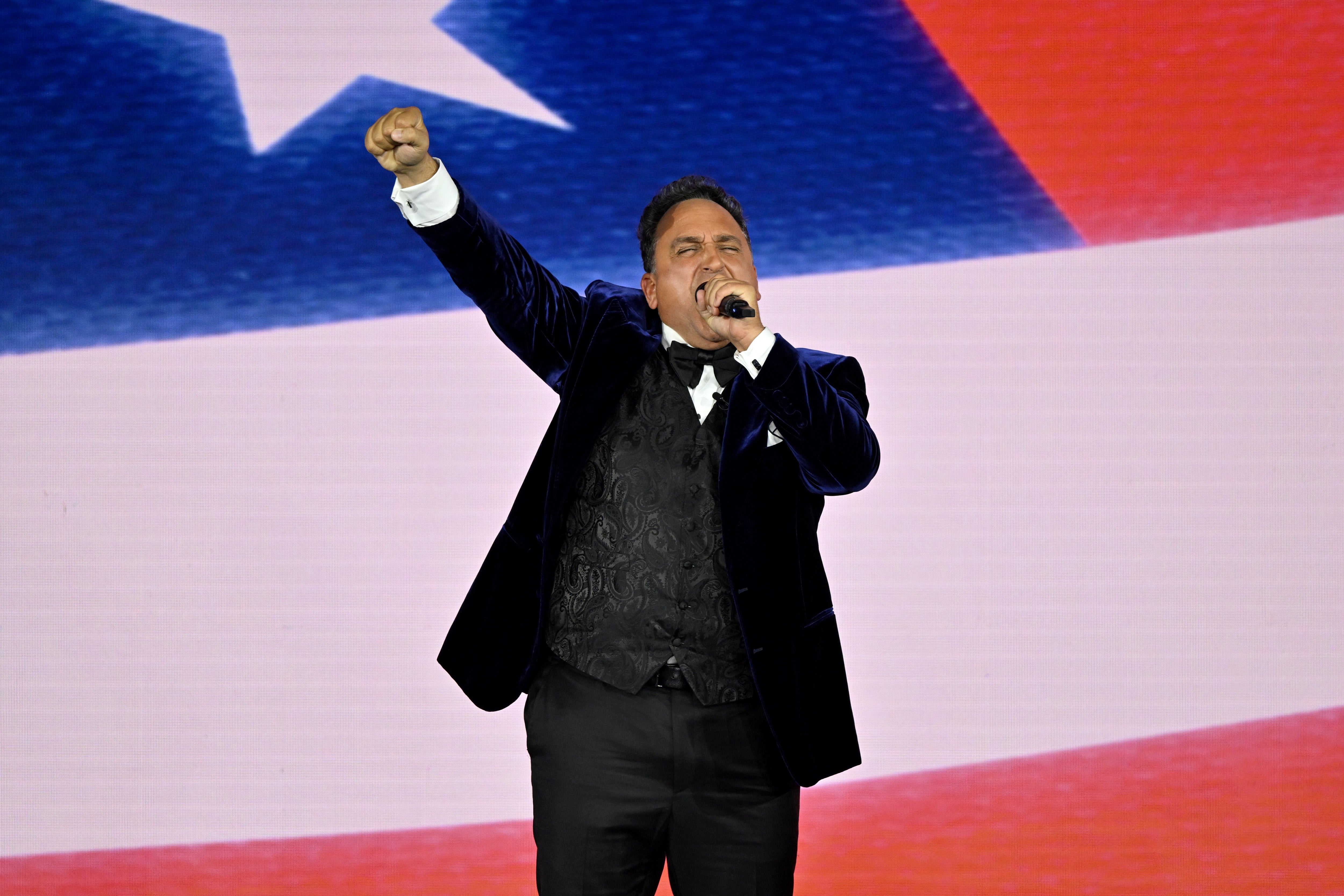 Person successful  a velvet suit   passionately sings connected  signifier    with a raised fist, against a backdrop resembling a flag