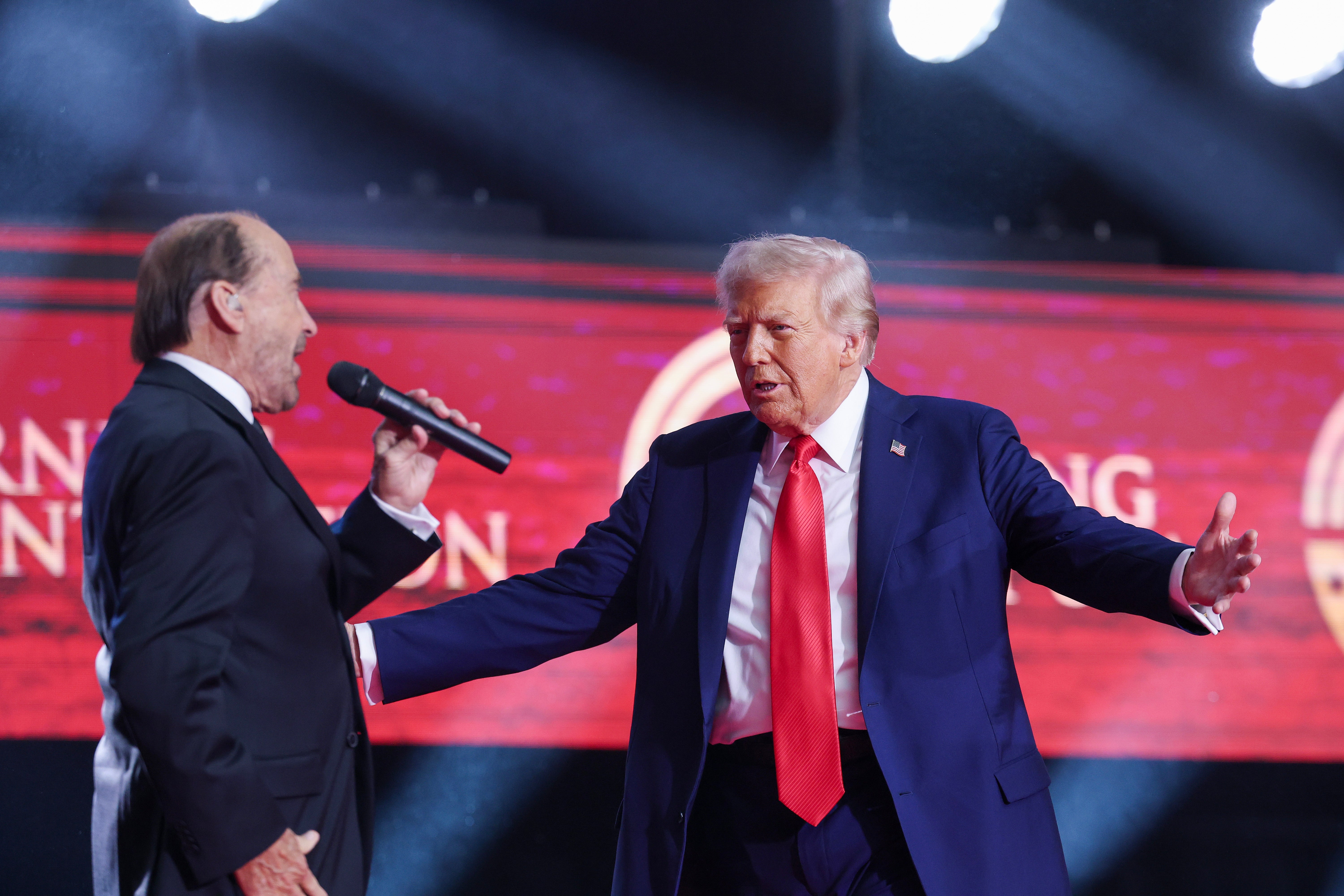 Two men connected  stage, 1  holding a microphone. Another, successful  a suit   with a agleam  reddish  tie, gestures animatedly