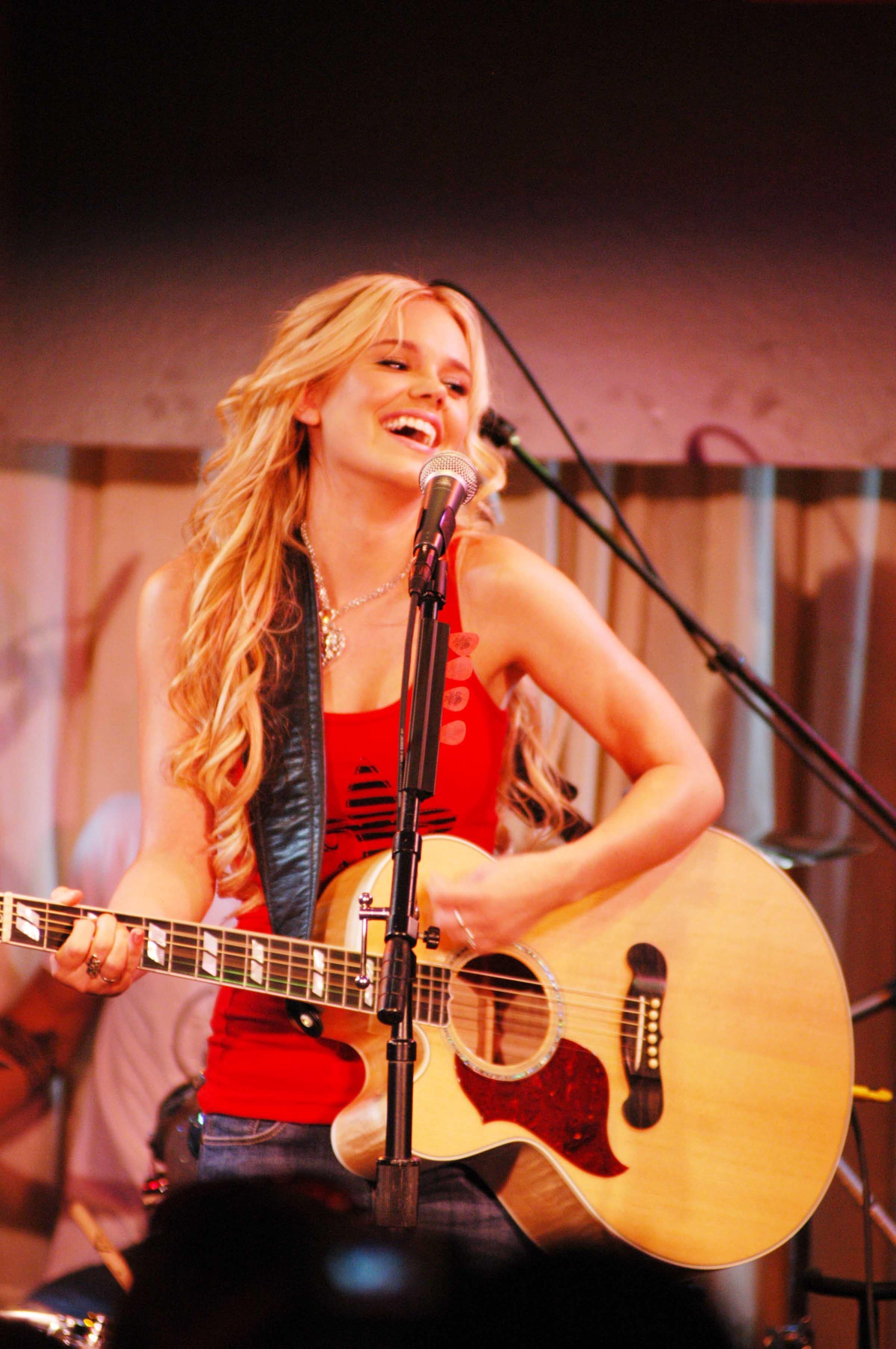Singer performs with a guitar on stage, smiling and engaging with the audience. She wears casual, stylish attire