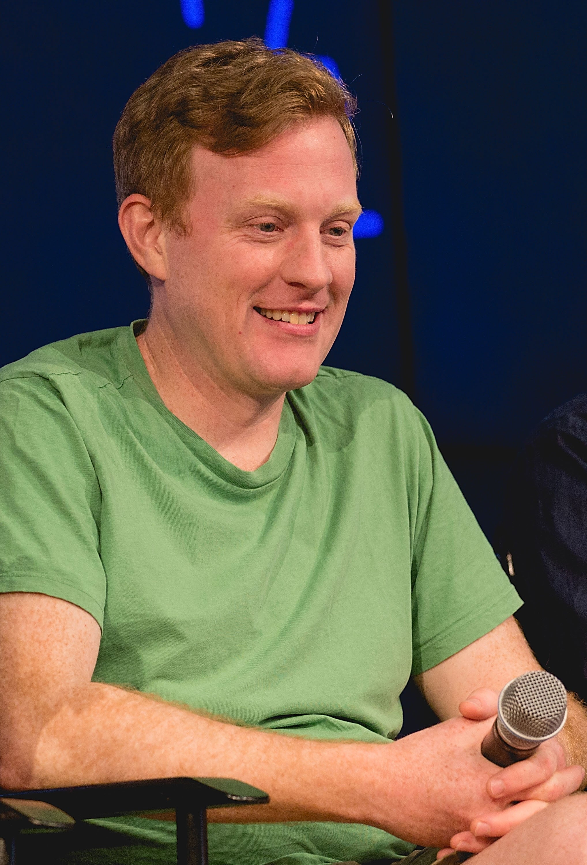 Person smiling while seated, wearing a casual top, holding a microphone at an event