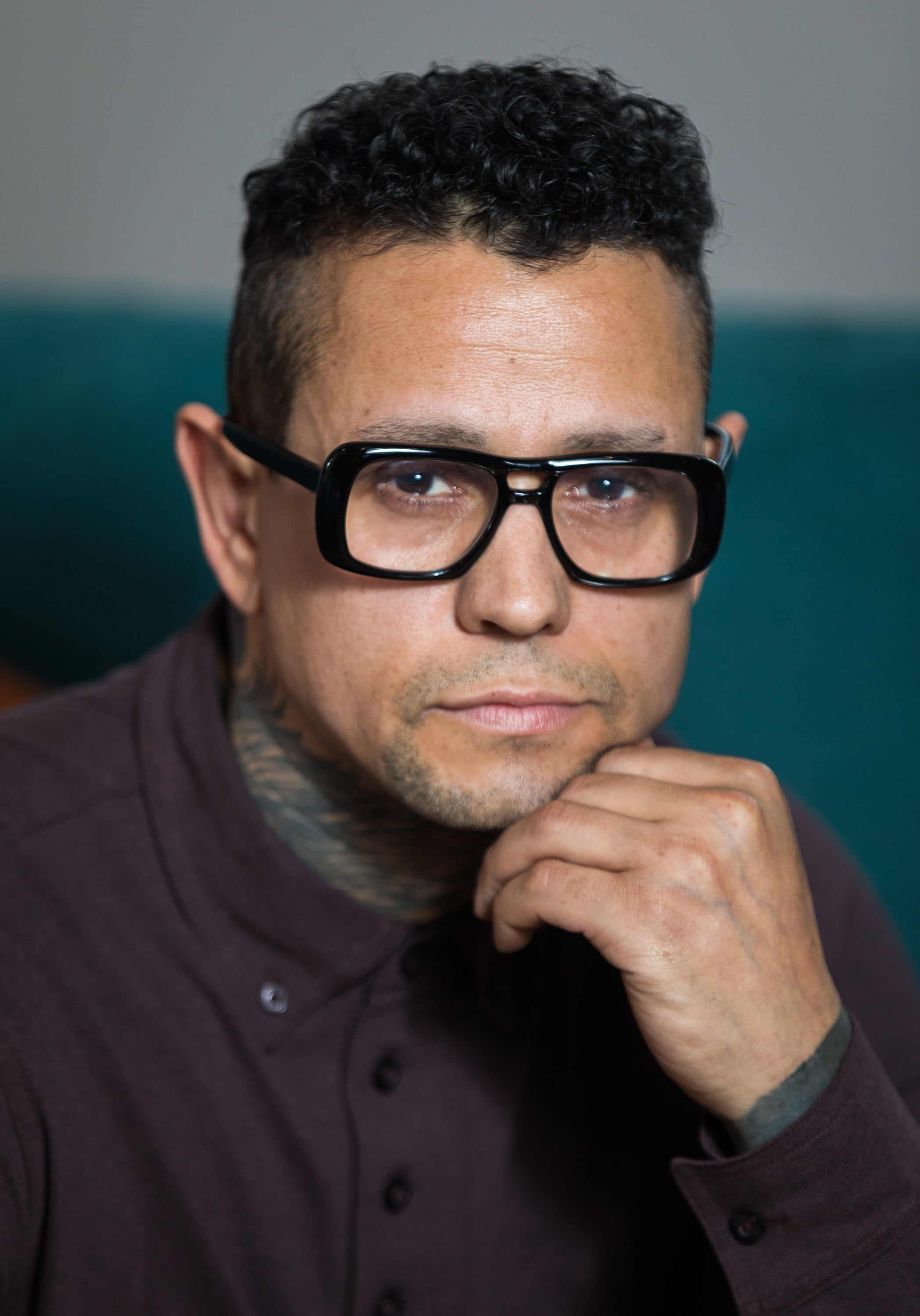 A person with short curly hair and large glasses, wearing a buttoned-up shirt, rests their chin on their hand, looking directly at the camera