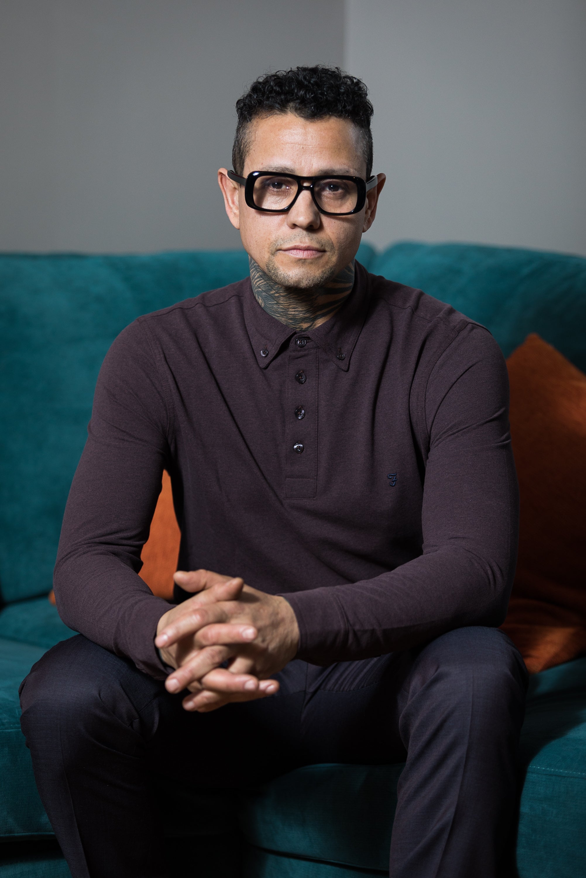 A person with glasses and tattoos sits on a sofa wearing a buttoned shirt, looking directly at the camera