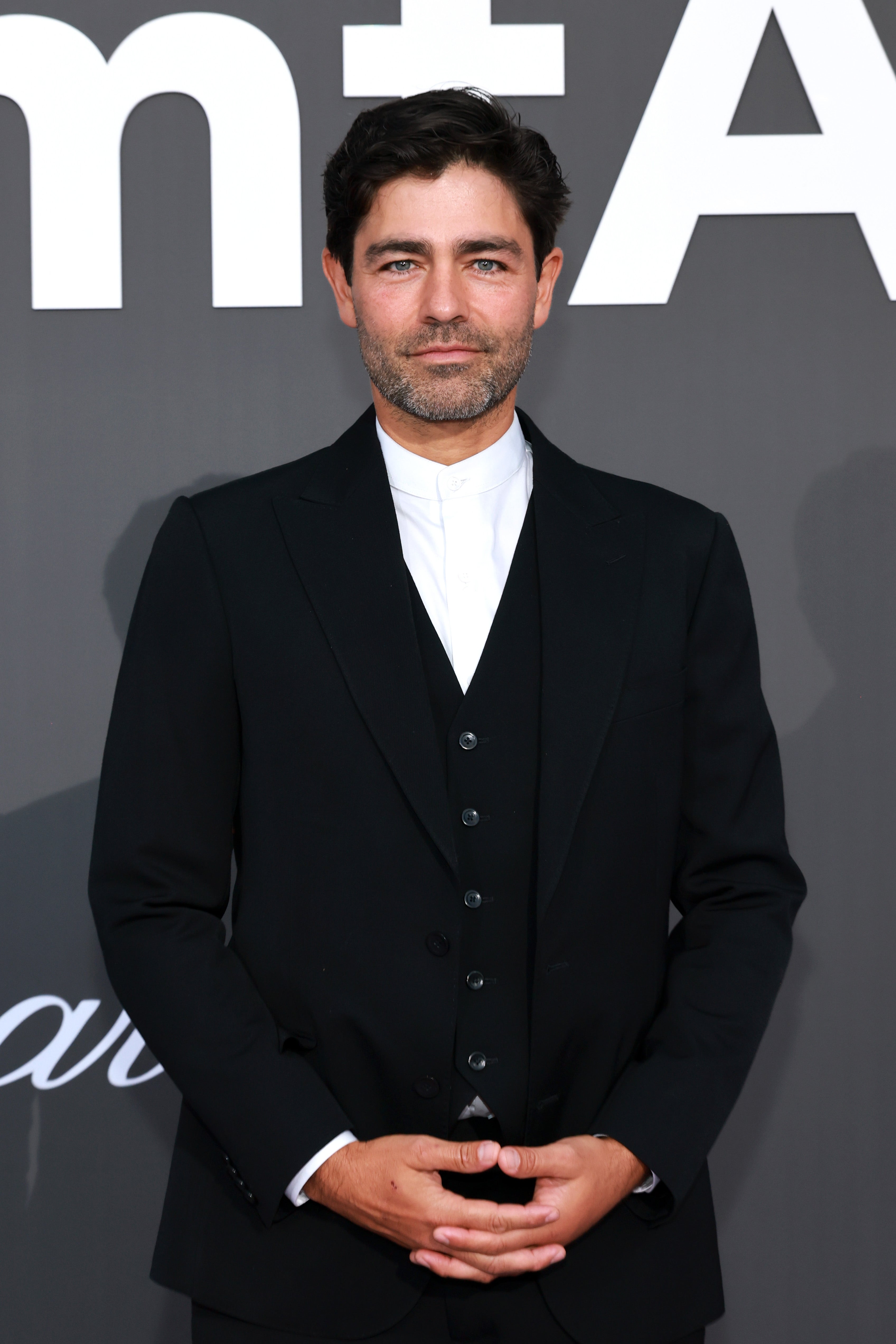 Man in a formal black suit with a white shirt stands in front of a media wall at a celebrity event