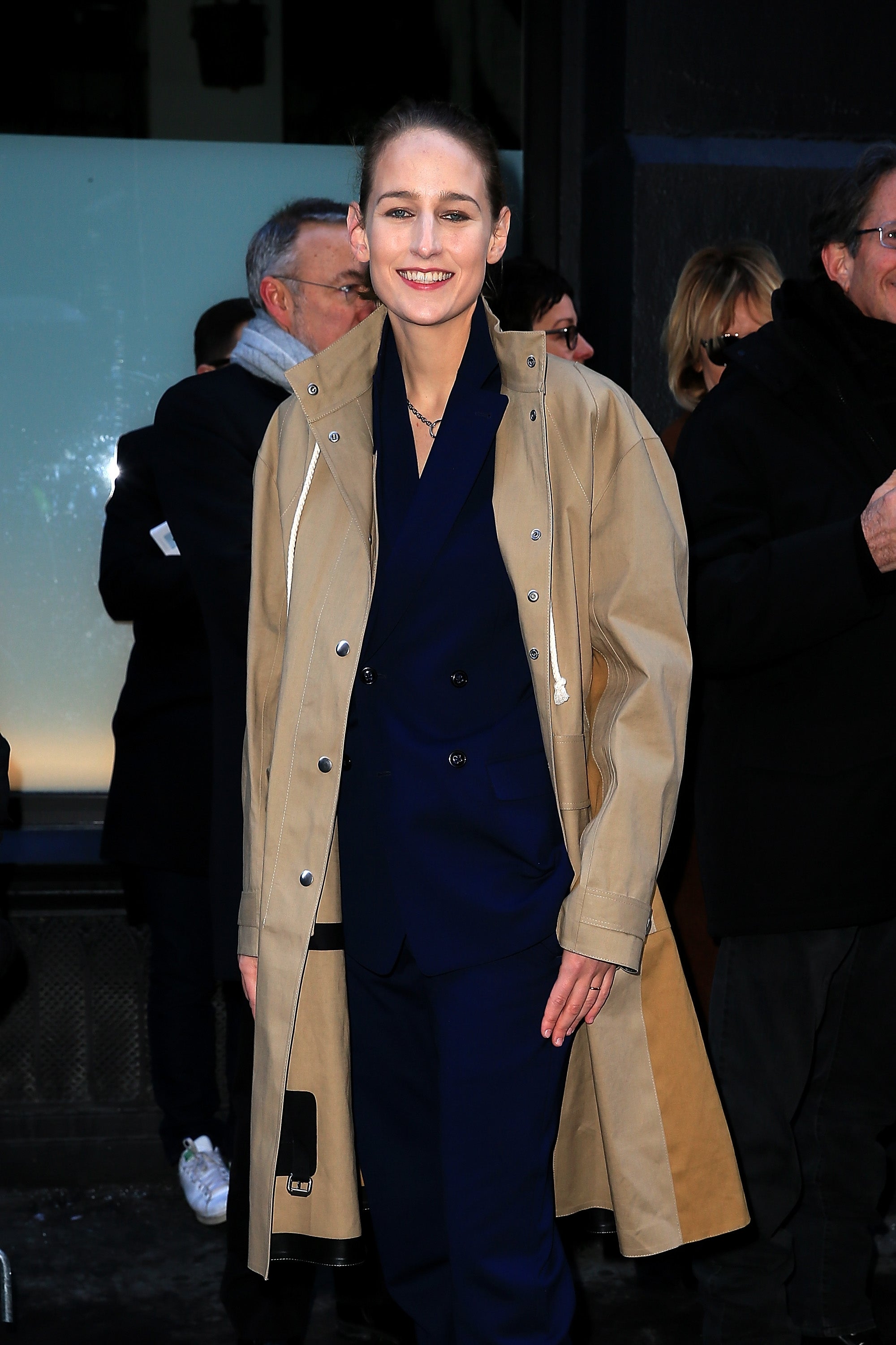 Person in a stylish blue suit and tan trench coat, smiling outdoors at a public event