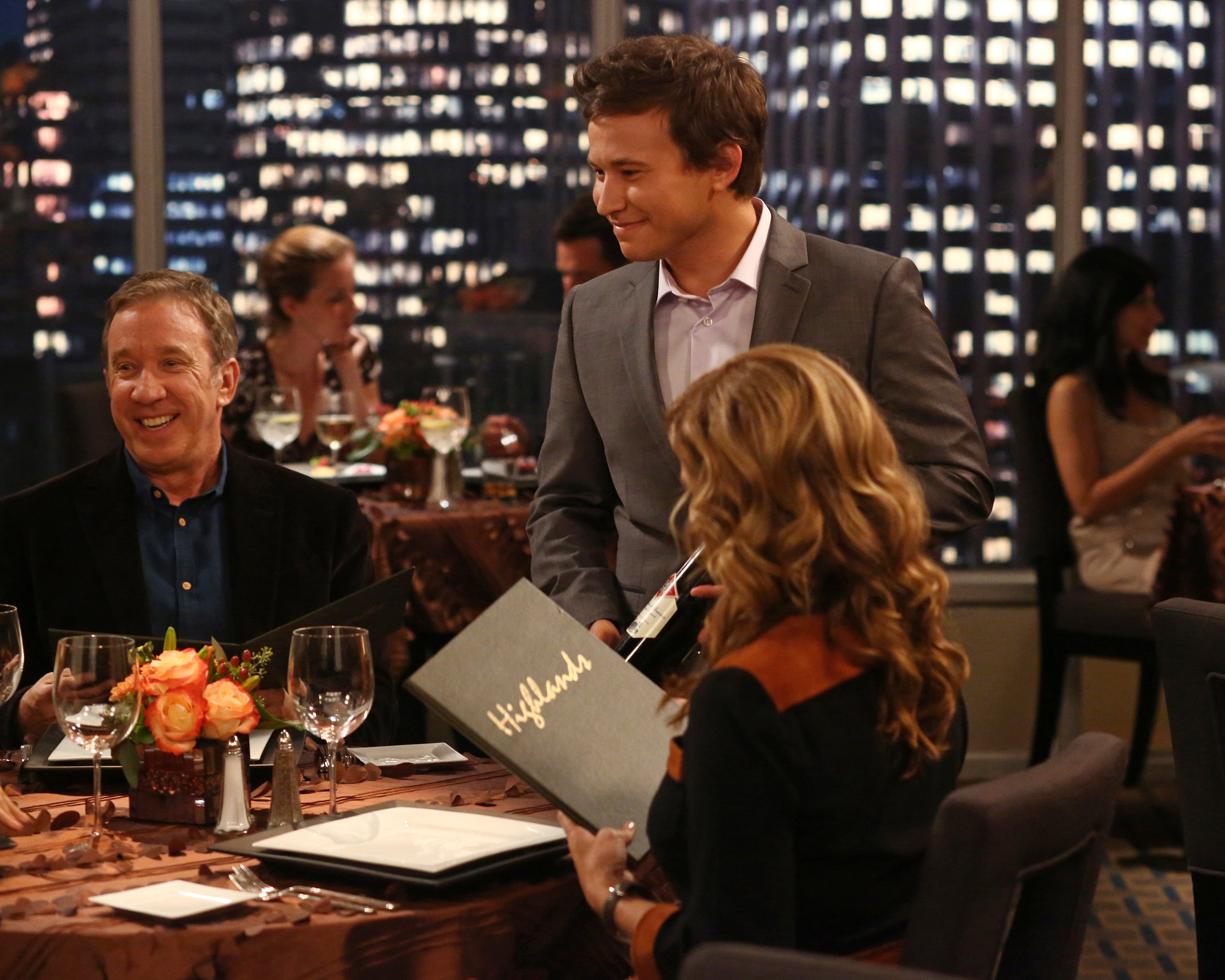 A group of people is dining at an elegant restaurant, with one person holding a menu. Urban cityscape is visible through large windows in the background