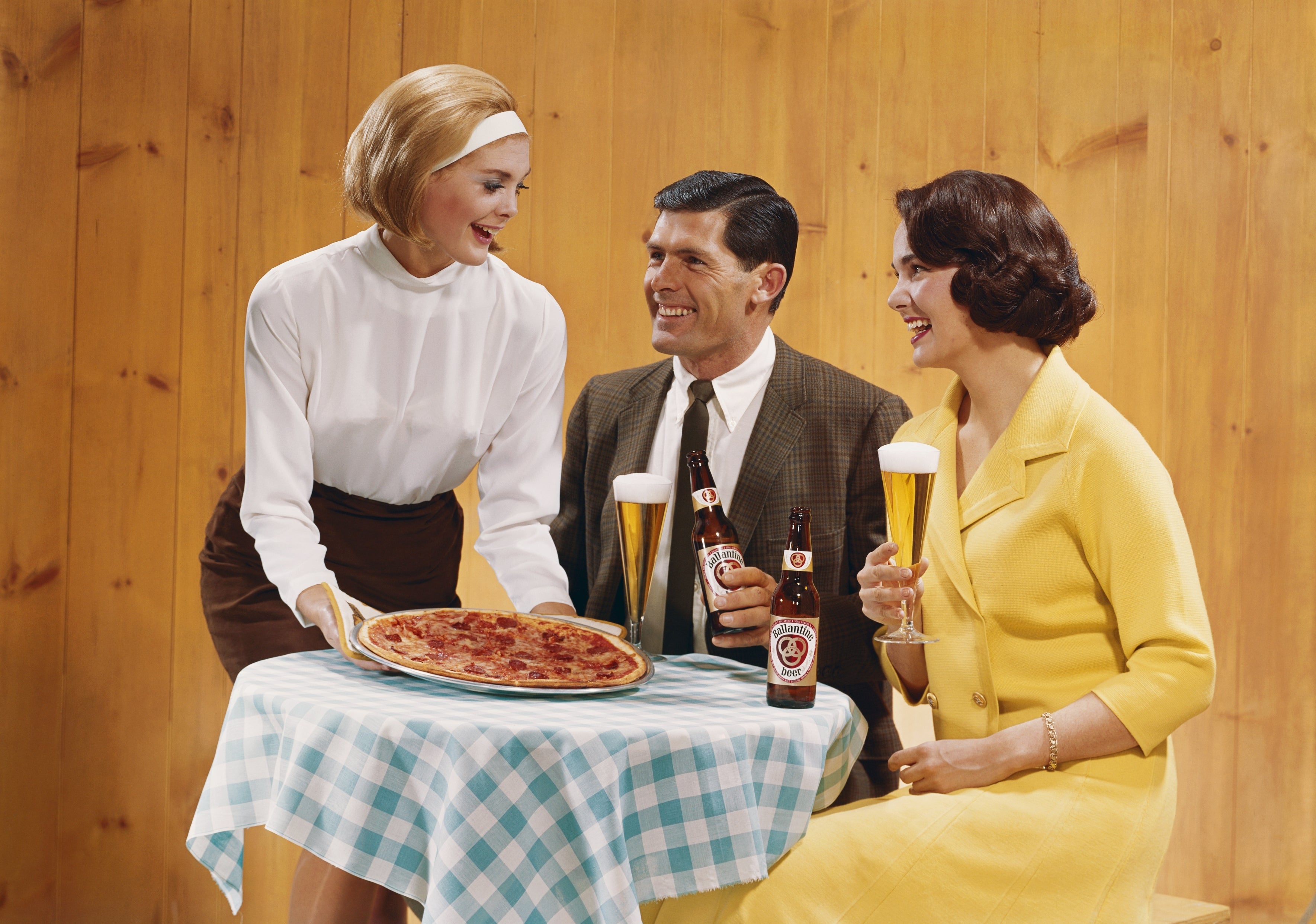 1960s-style country with 2 seated individuals enjoying pizza and beer, served by a smiling waitress. Retro manner and eating setting