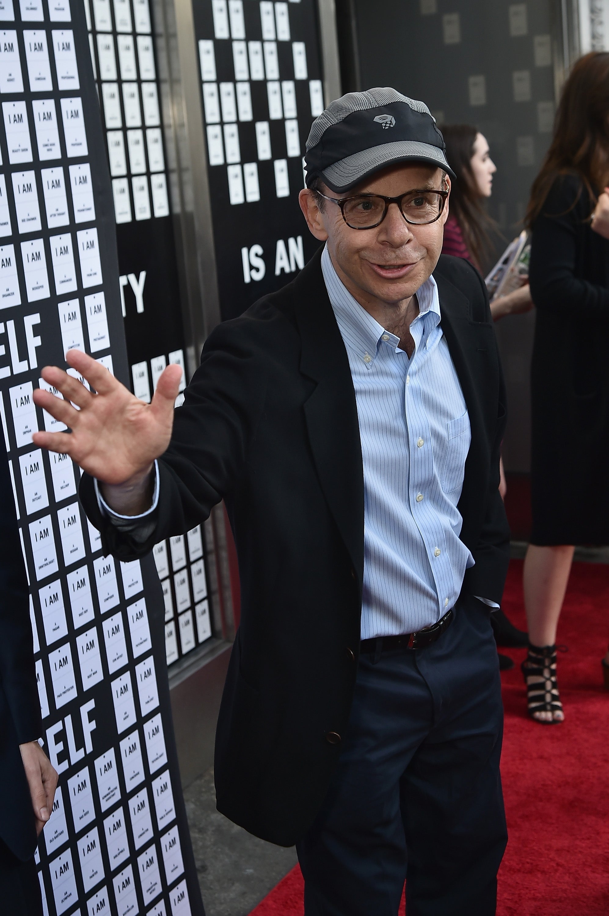 A person wearing a blazer, striped shirt, and cap waves on the red carpet at an event with text-covered walls