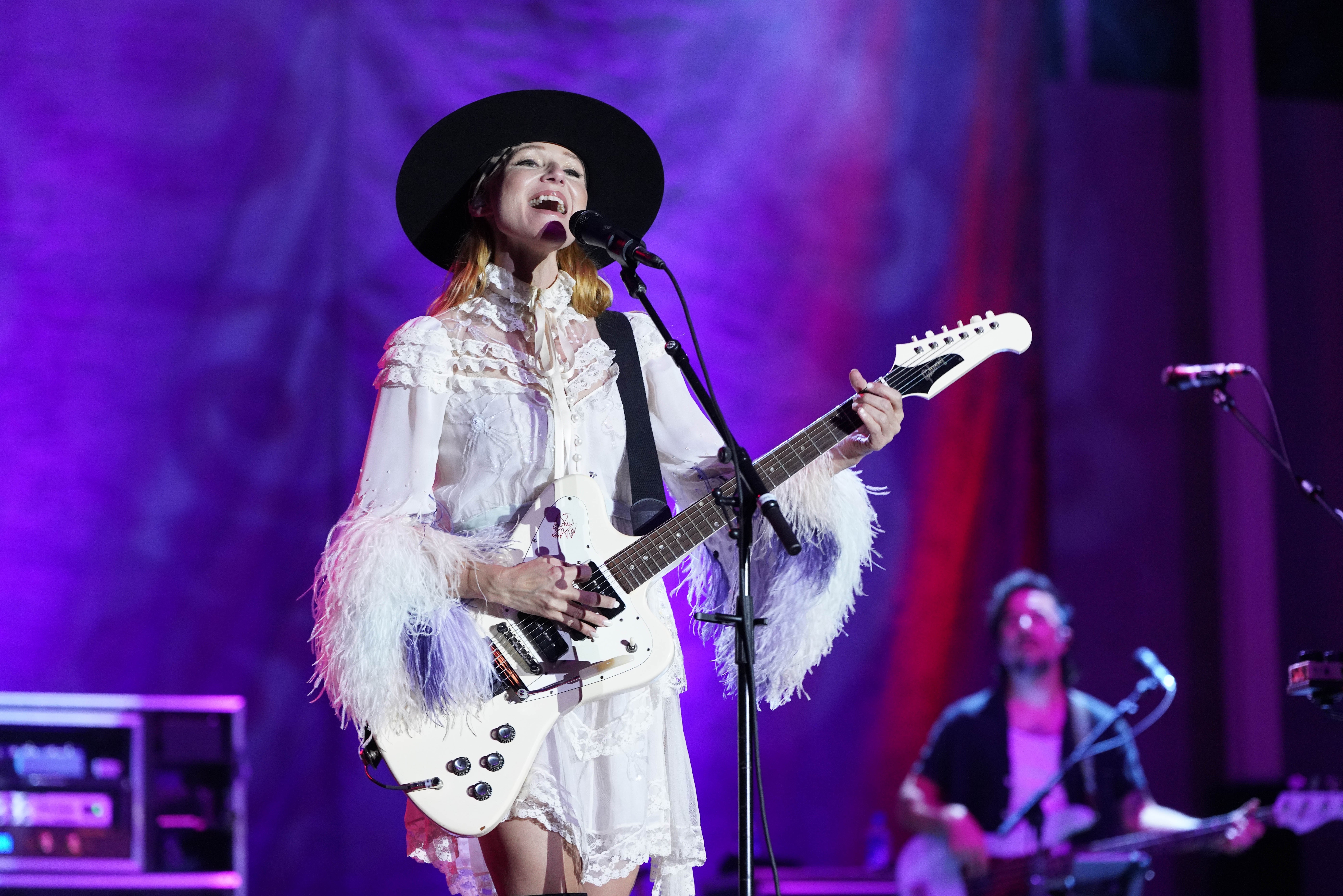 Musician on stage wearing a fringed dress and wide-brim hat, performing with an electric guitar