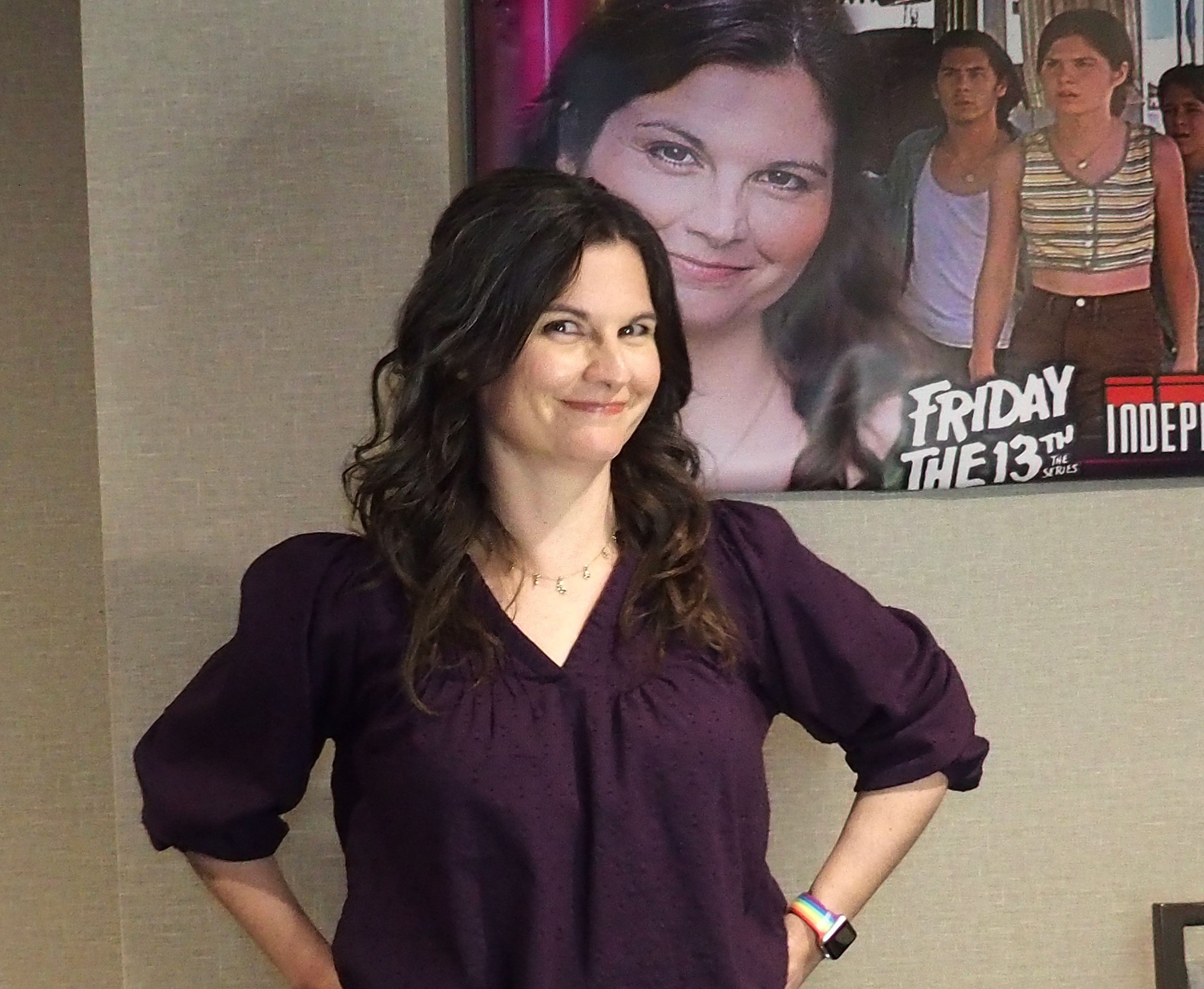 Person smiling in front of a "Friday the 13th" poster, wearing a casual purple blouse with wavy hair