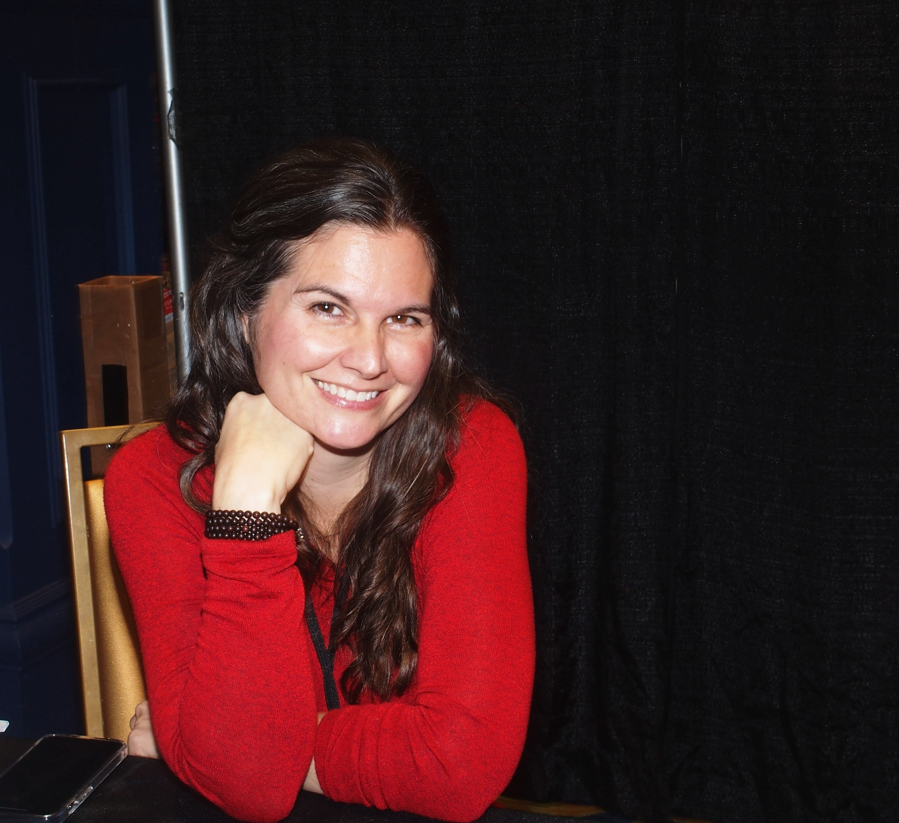 Smiling woman with long hair in a relaxed pose, wearing a casual red long-sleeve top, seated at a table against a dark backdrop