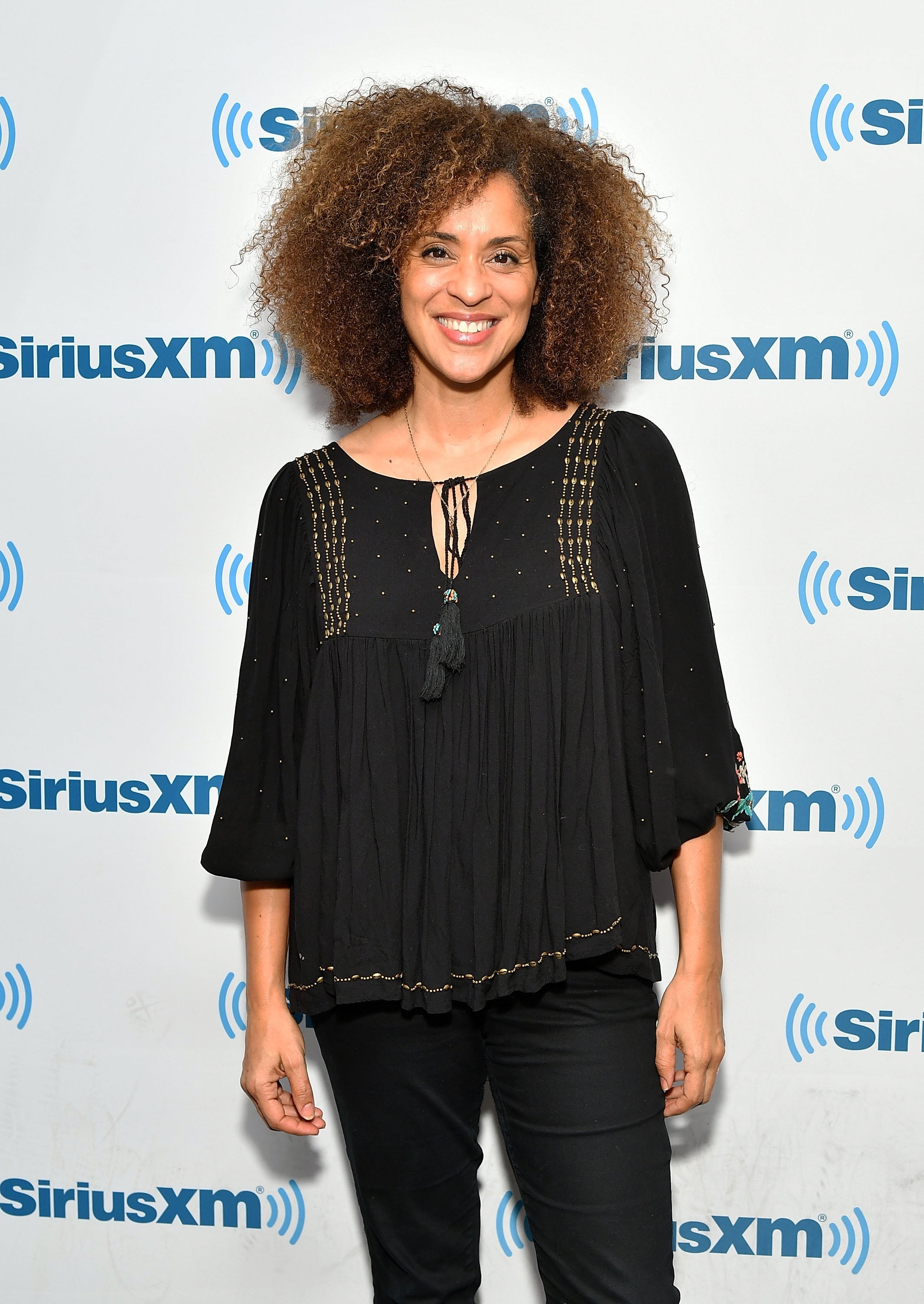 A person with curly hair smiles in a flowy, black top with gold accents at a media event with a SiriusXM backdrop