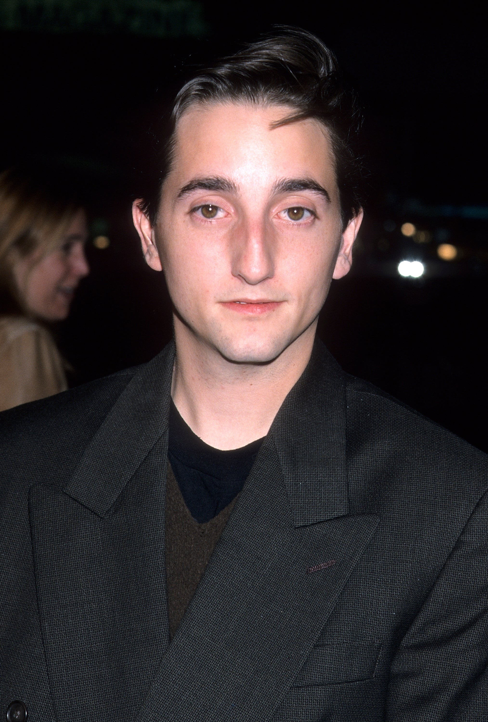 A person in a formal suit at an event, posing for the camera with a neutral expression. Another person is visible in the background