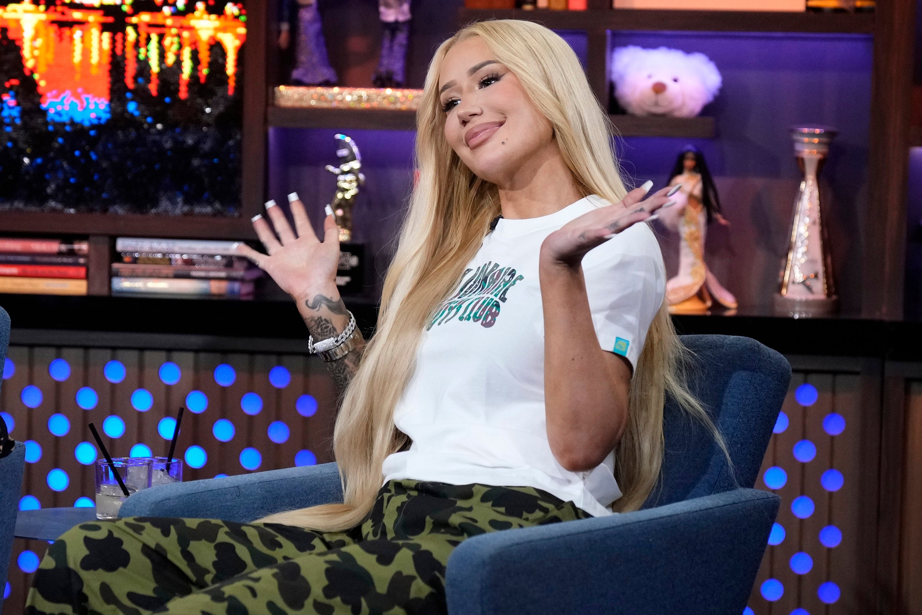 Celebrity wearing a casual white T-shirt and camouflage pants, seated with a playful shrug on a talk show set