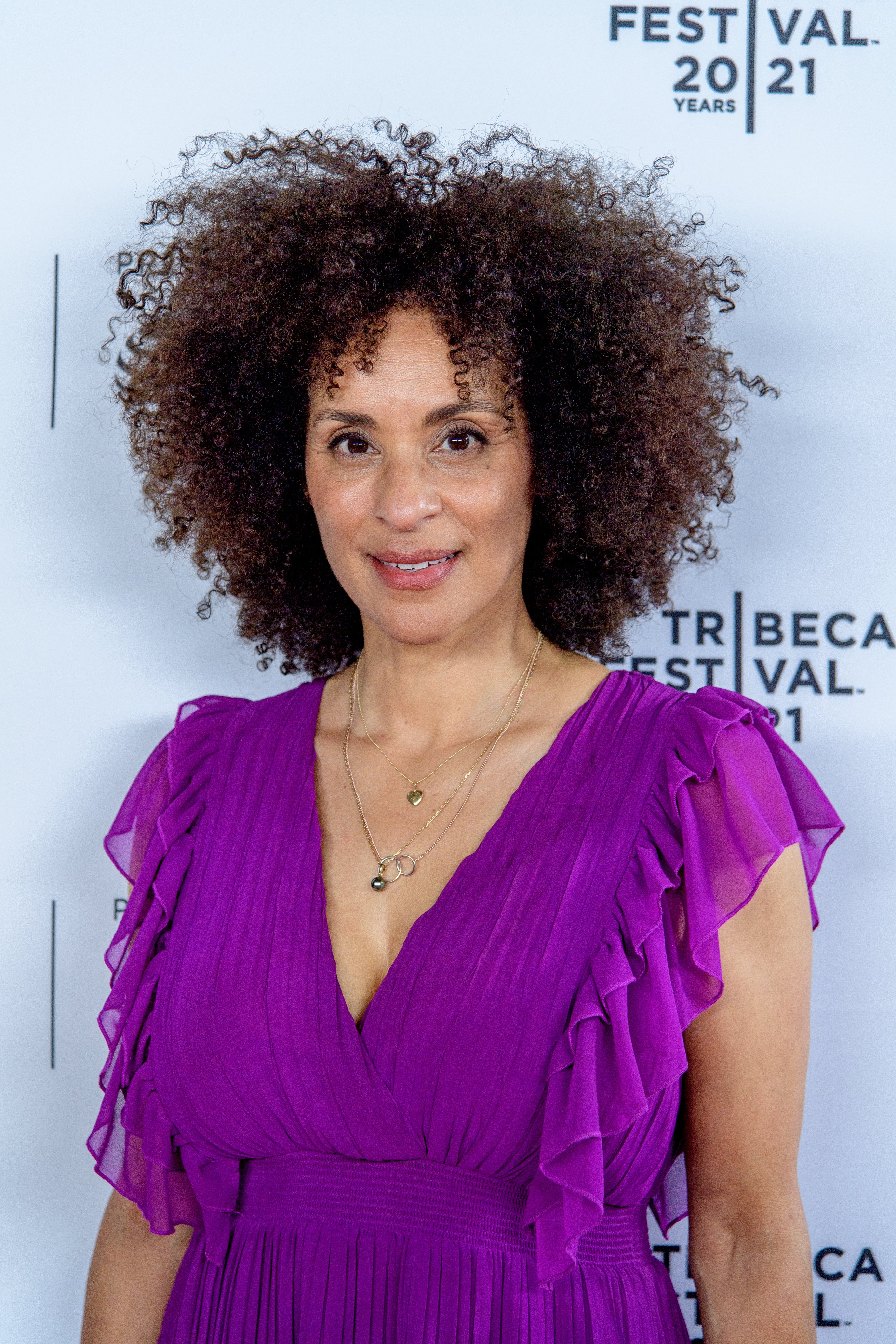 Person at an event wears a ruffled dress with layered necklaces, standing in front of a backdrop with "Tribeca Festival" text