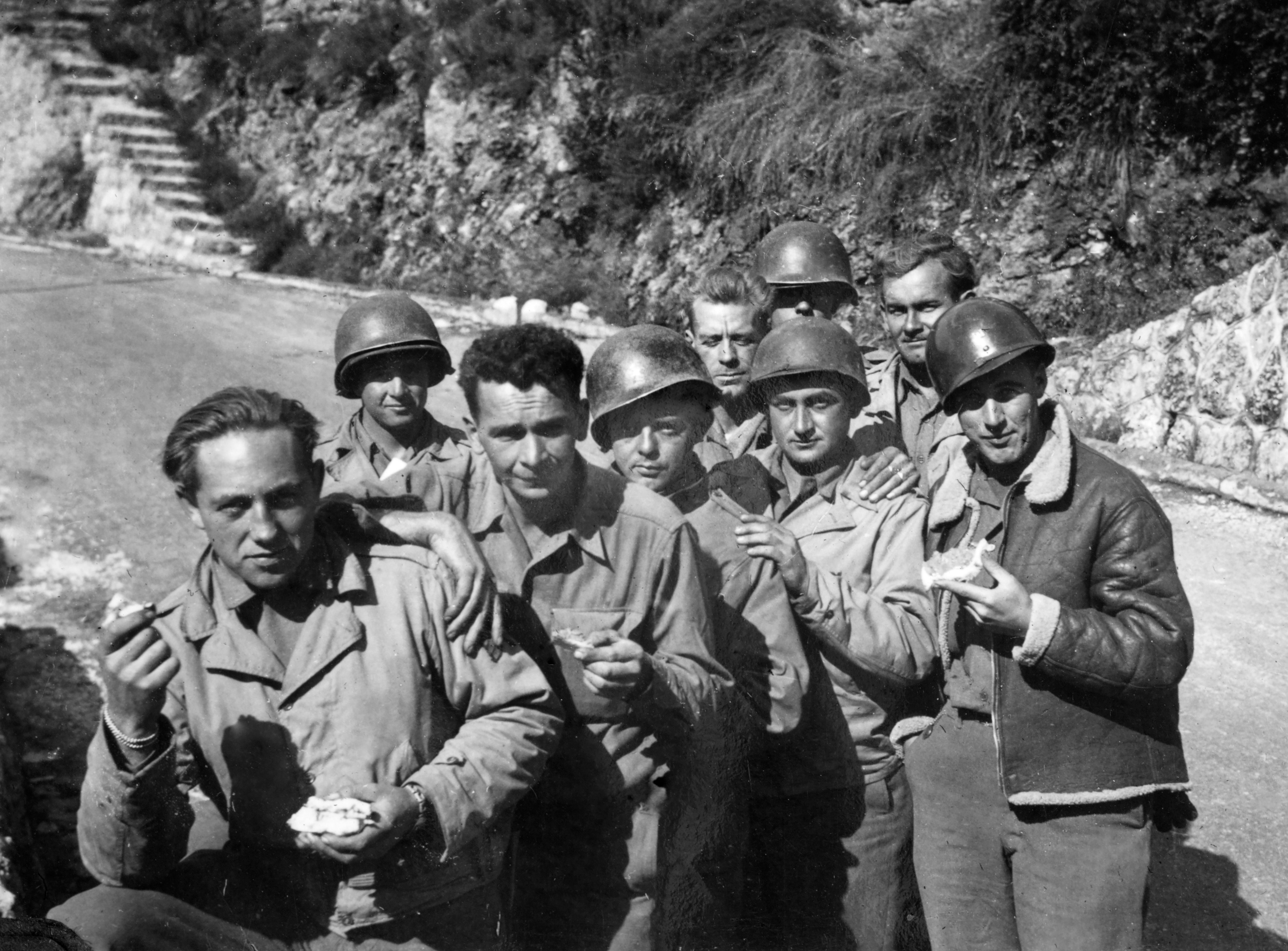 A radical of soldiers stands connected a roadside, wearing helmets and casual subject attire, immoderate holding food, with rocky terrain disposable successful the background