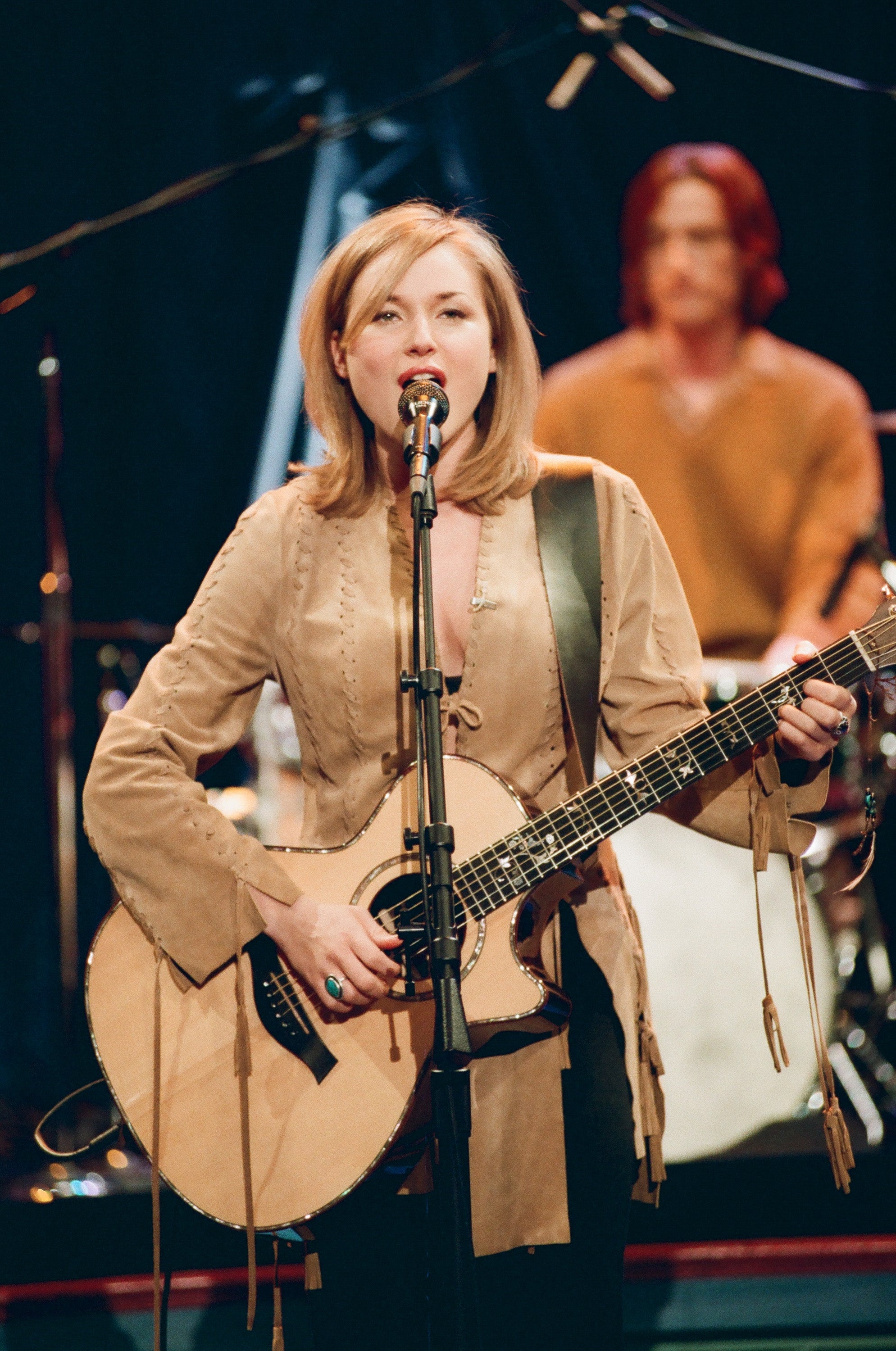 Musician performing on stage, playing an acoustic guitar and singing into a microphone, with a drummer in the background