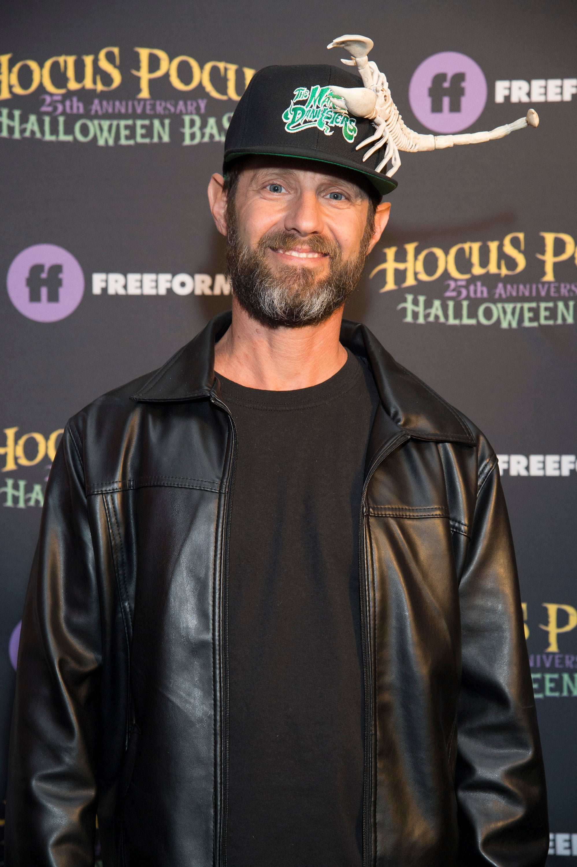 A man on a red carpet wears a leather jacket and a cap with a 3D scorpion design at a "Hocus Pocus 25th Anniversary Halloween" event